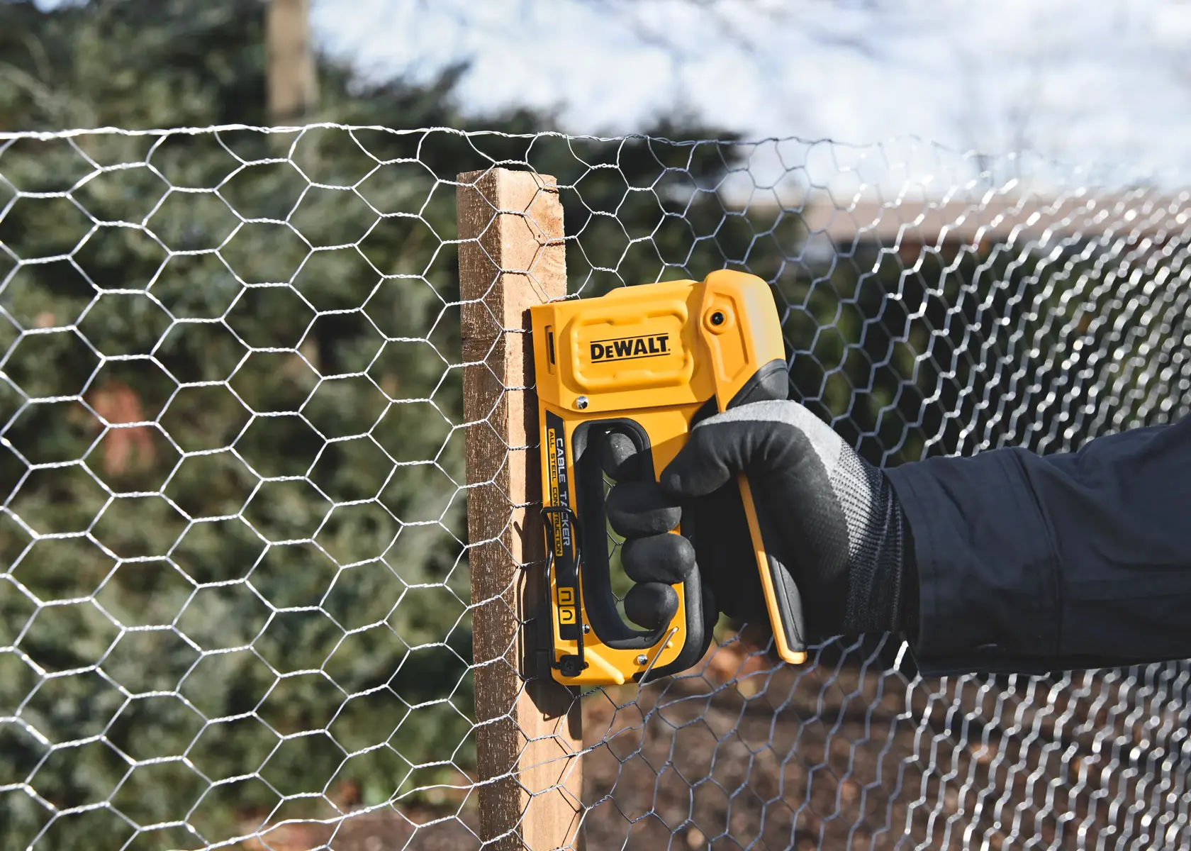 A person wearing a black glove uses a yellow DEWALT staple gun to secure hexagonal wire mesh to a wooden post outdoors.