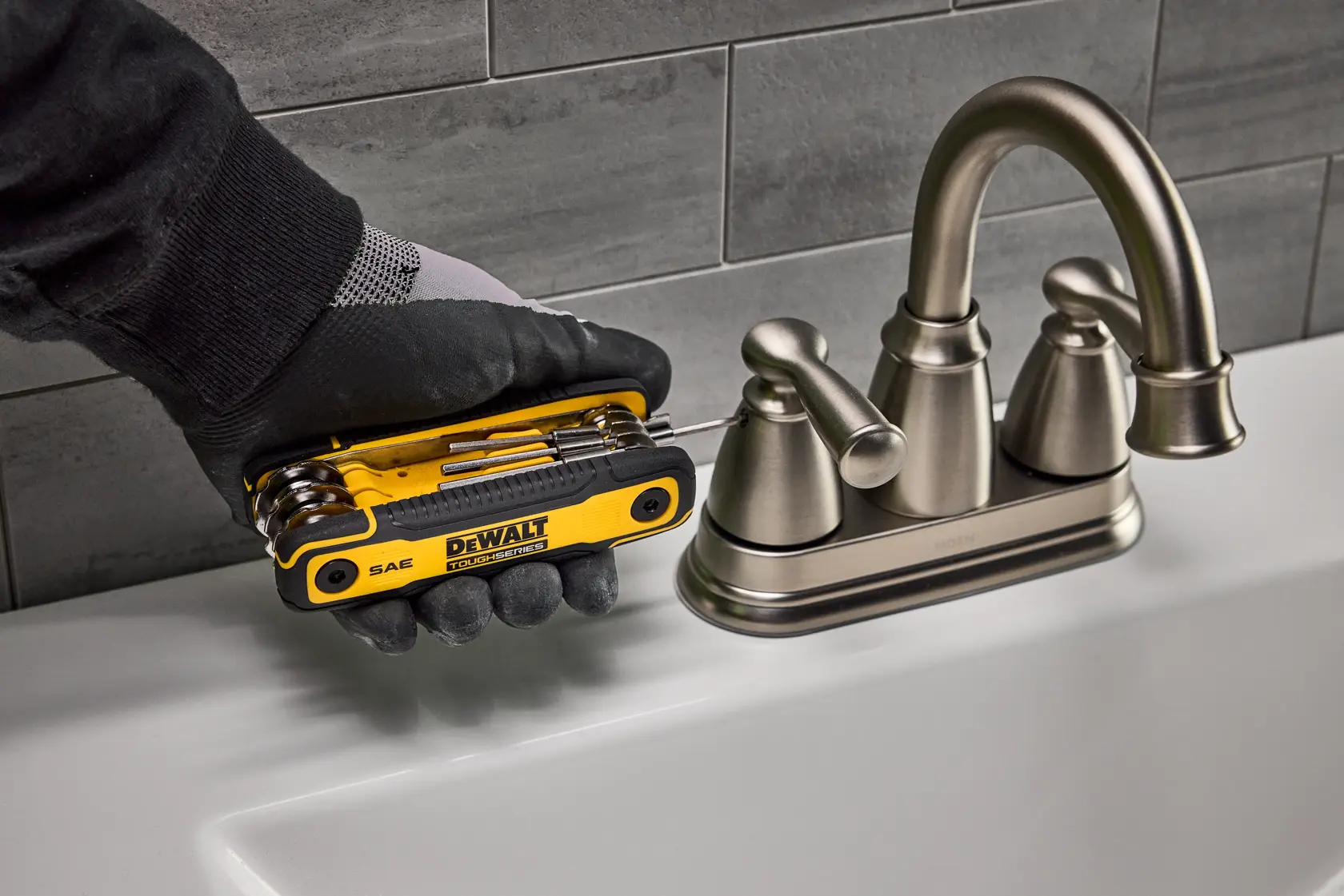 A person wearing a black glove uses a DEWALT DWHT70266 multi-tool to adjust a silver bathroom faucet. The multi-tool is yellow and black and has multiple folding tools visible.