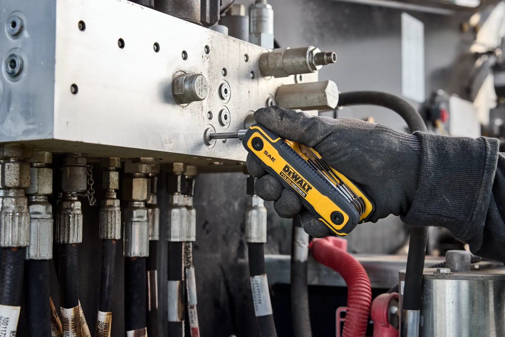 Gloved hand uses a DEWALT folding hex key tool on a metal hydraulic machine with hoses and connectors.