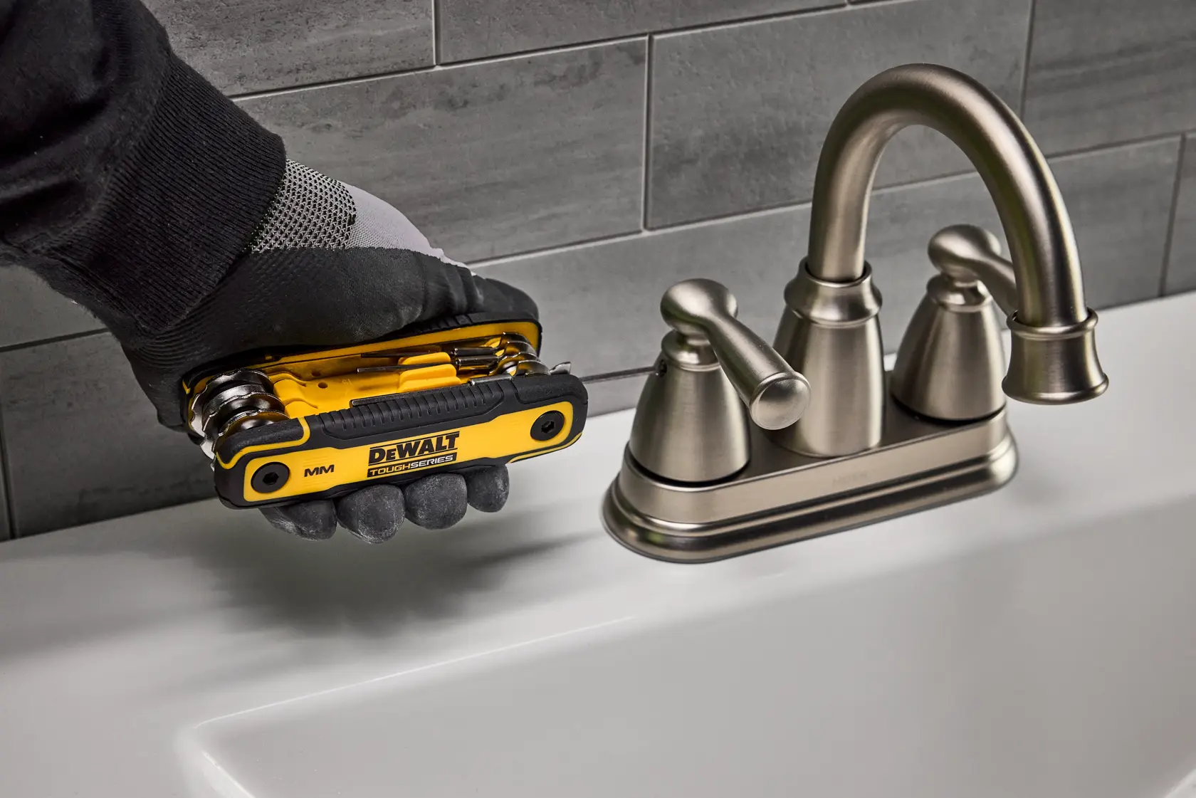 A gloved hand holds a DEWALT folding hex key tool near a metal bathroom faucet on a white sink, with gray tiled wall in the background.