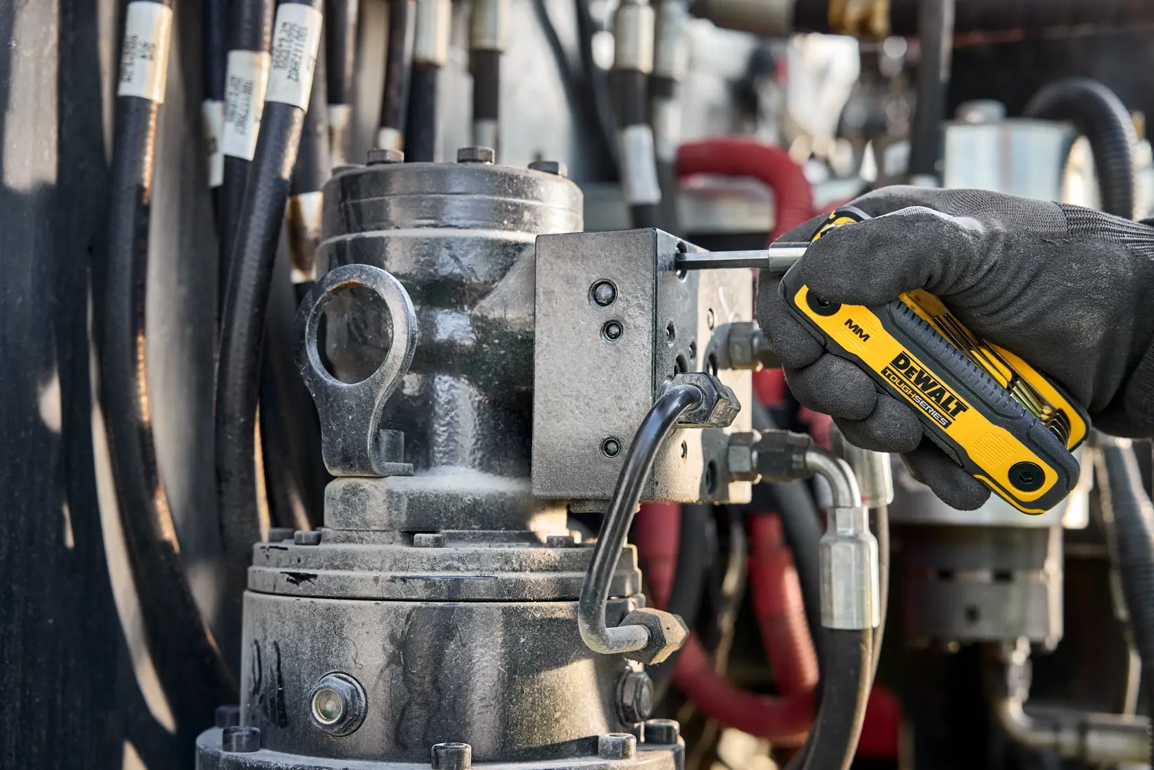 A gloved hand uses a DEWALT folding hex key tool to adjust a metal component with connected hoses in an industrial setting.