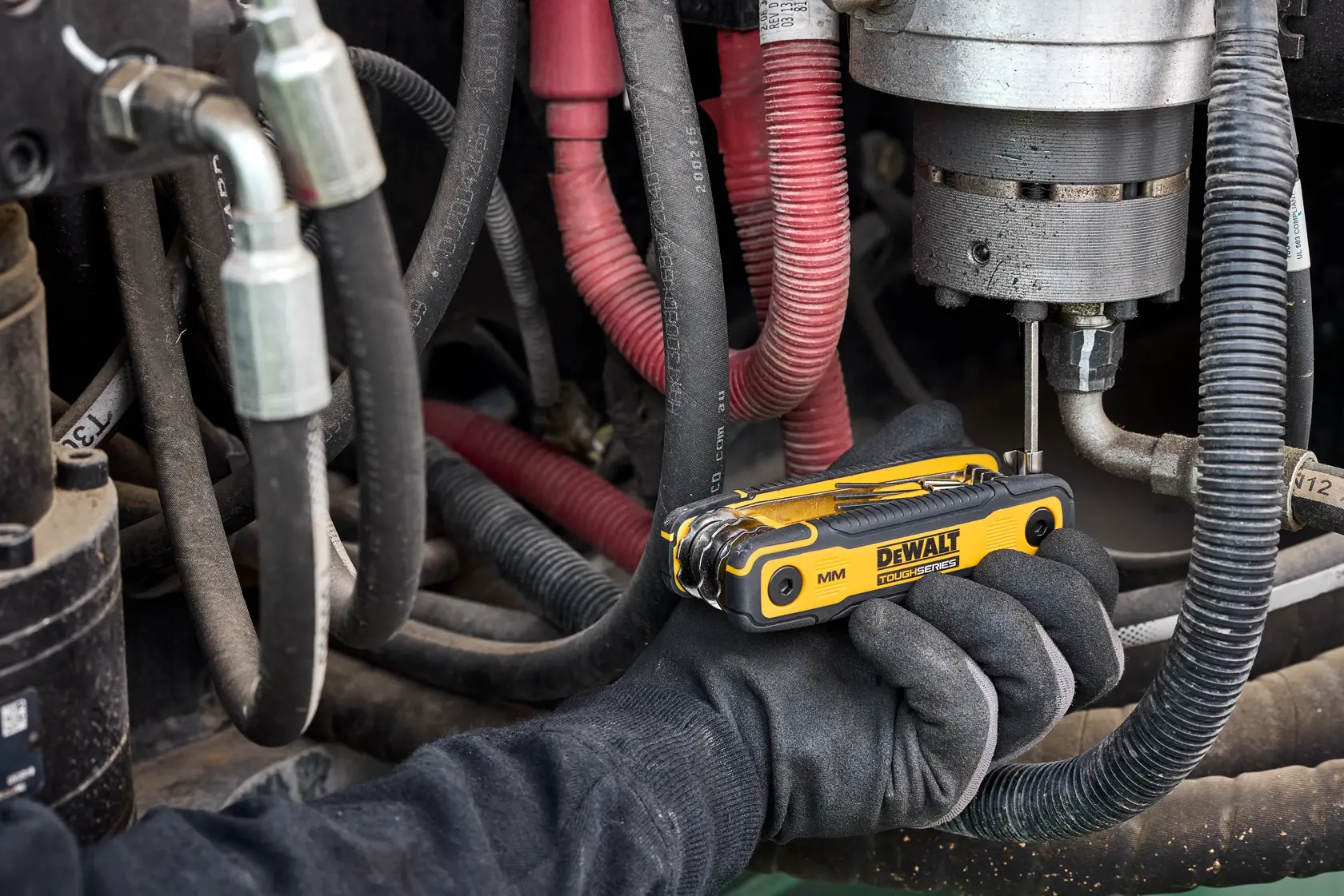 A hand wearing a black glove uses a yellow DEWALT multi-tool to work on machinery with several cables and hoses.