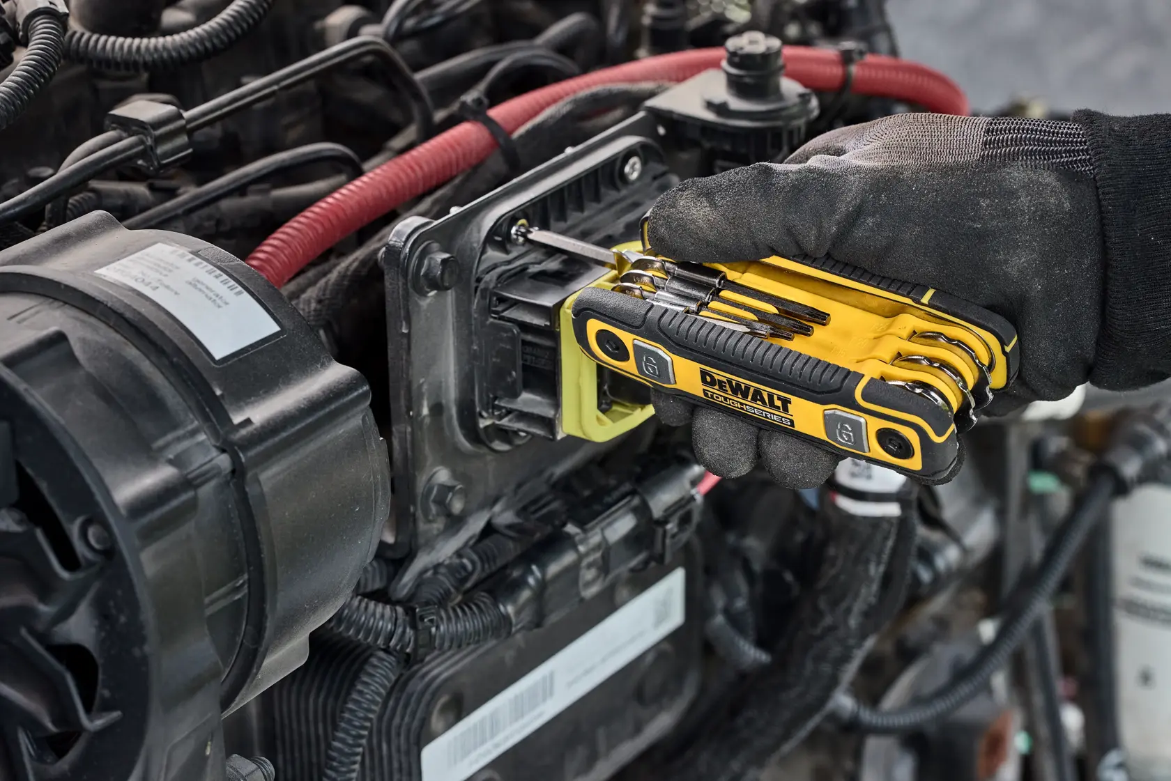 A gloved hand uses a DEWALT folding hex key set to adjust or tighten a bolt on a mechanical engine component, surrounded by wires and parts.