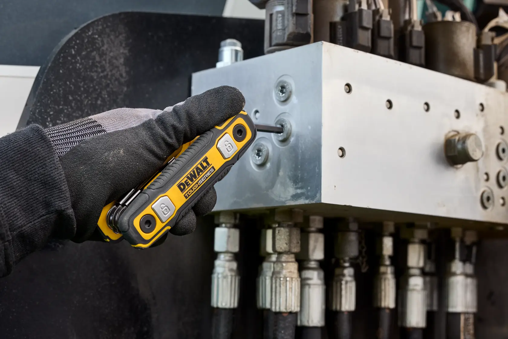 A gloved hand uses a DEWALT folding hex key tool to adjust a metal hydraulic component with multiple hoses and fittings.