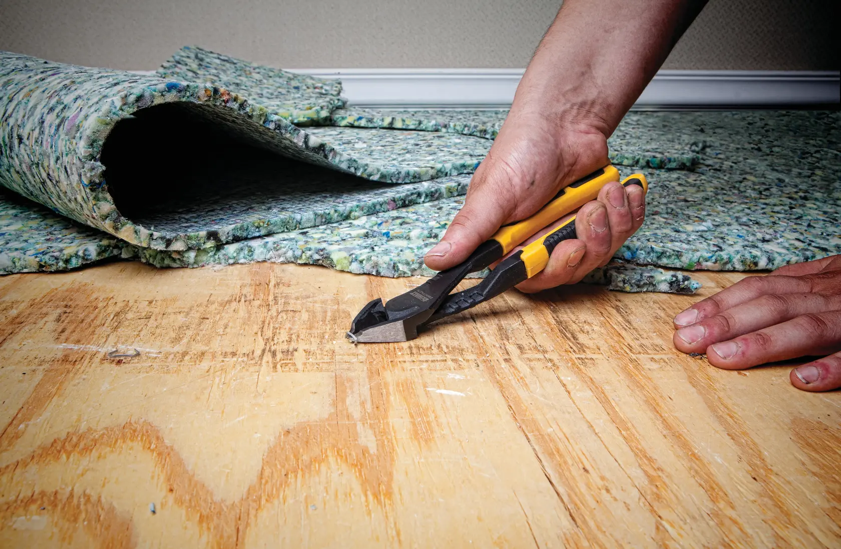 A close-up of a person's hand using a DEWALT yellow and black diagonal cutting plier to cut a staple from plywood flooring, with carpet padding partially lifted in the background.
