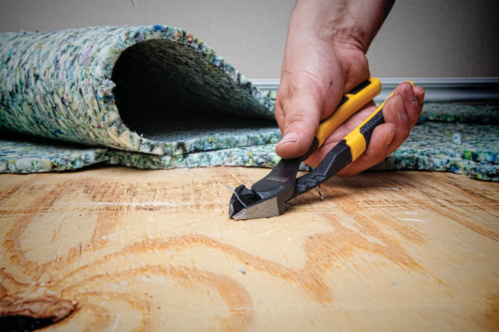 A hand is holding a DEWALT cutting tool with yellow and black handles, using it to cut or pull something from a wooden floor. In the background, there is a rolled-up piece of carpet padding.
