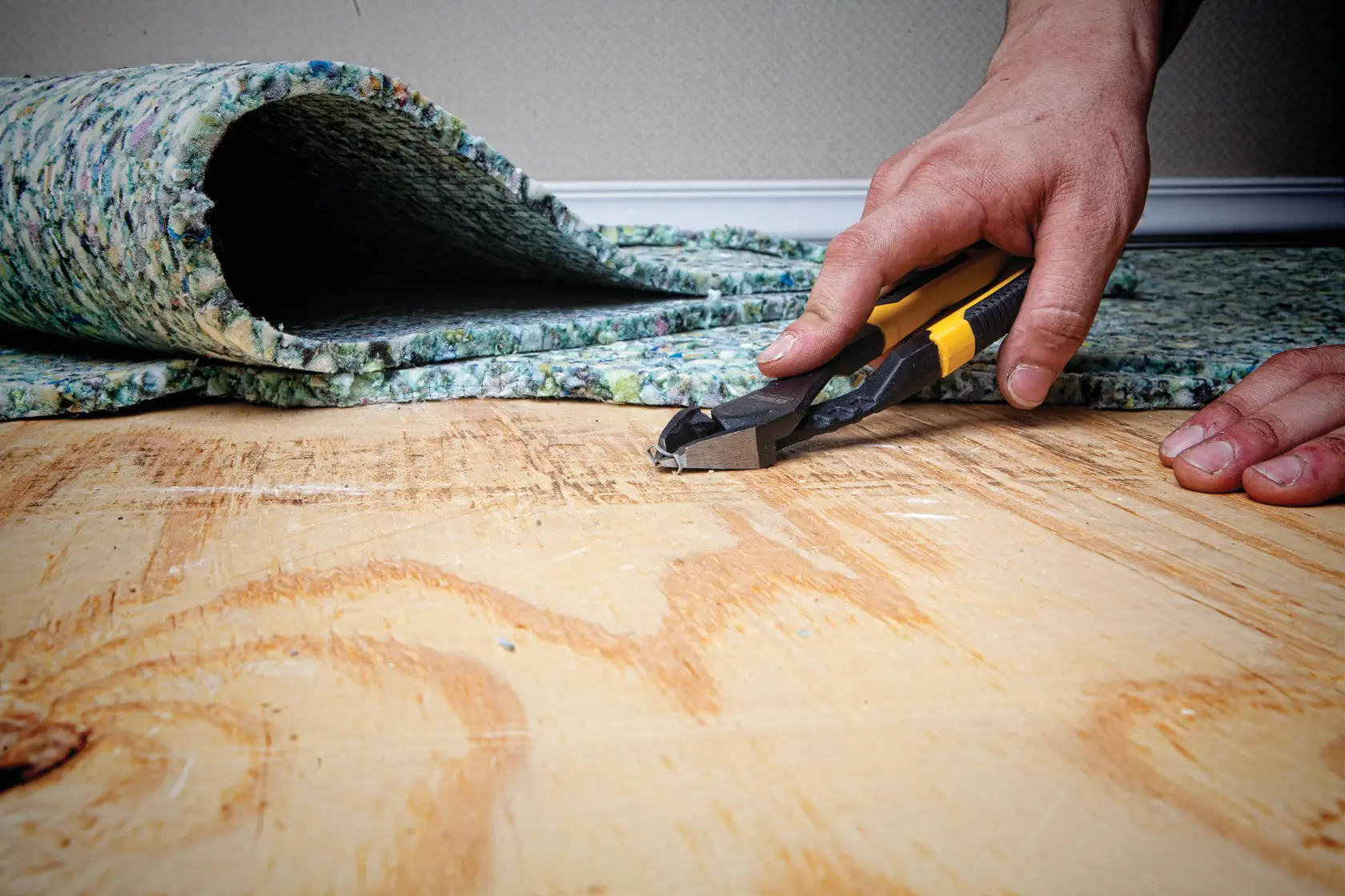 A close-up of a person's hand using a DEWALT cutting tool (DWHT74274) to remove a staple from a wooden floor, with carpet padding partially lifted in the background.