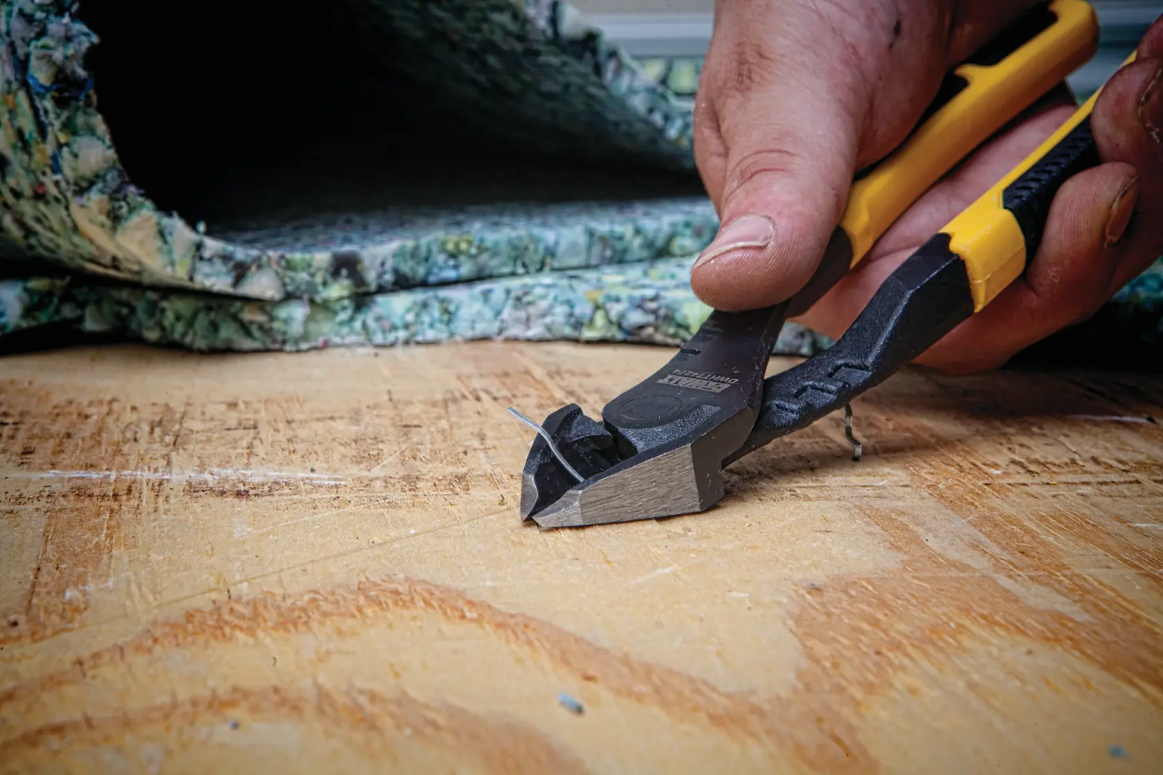 A close-up of a hand using DEWALT diagonal cutting pliers to cut a small wire on a wooden surface, with carpet padding in the background.