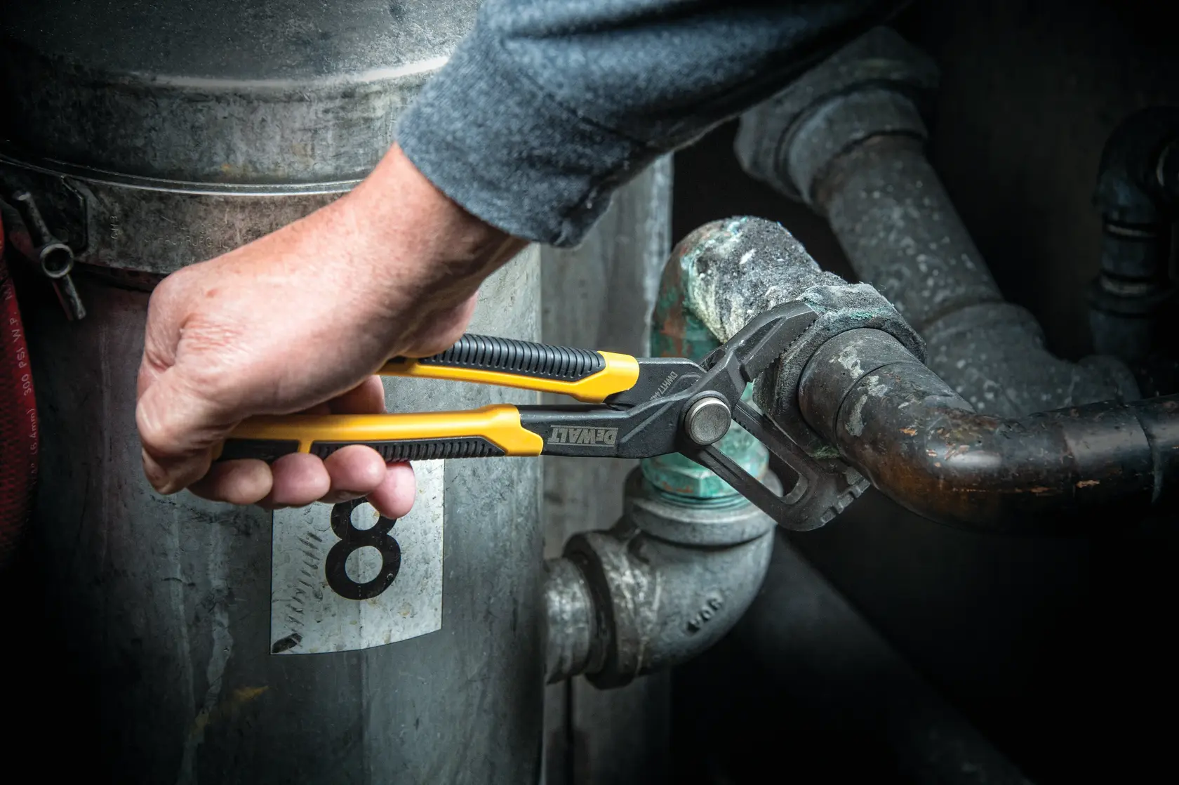 A person uses a DEWALT adjustable groove joint plier to grip and turn a metal pipe in an industrial setting.