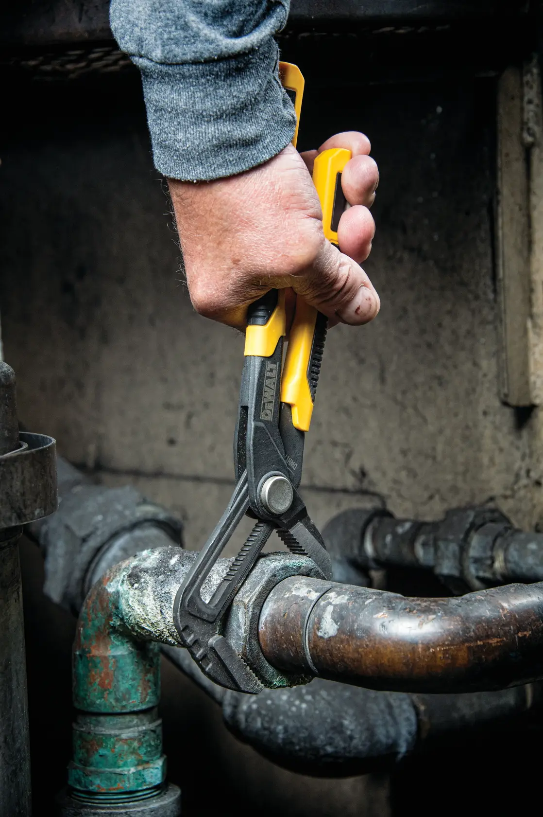 A person is using a DEWALT adjustable plier to grip and turn a corroded metal pipe fitting in an industrial setting.