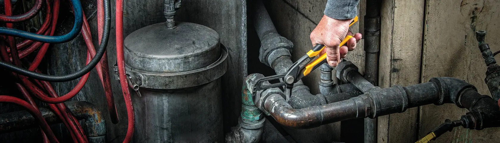 A person uses a DEWALT adjustable wrench to tighten or loosen a metal pipe fitting in an industrial setting. Several pipes and hoses are visible around the workspace.