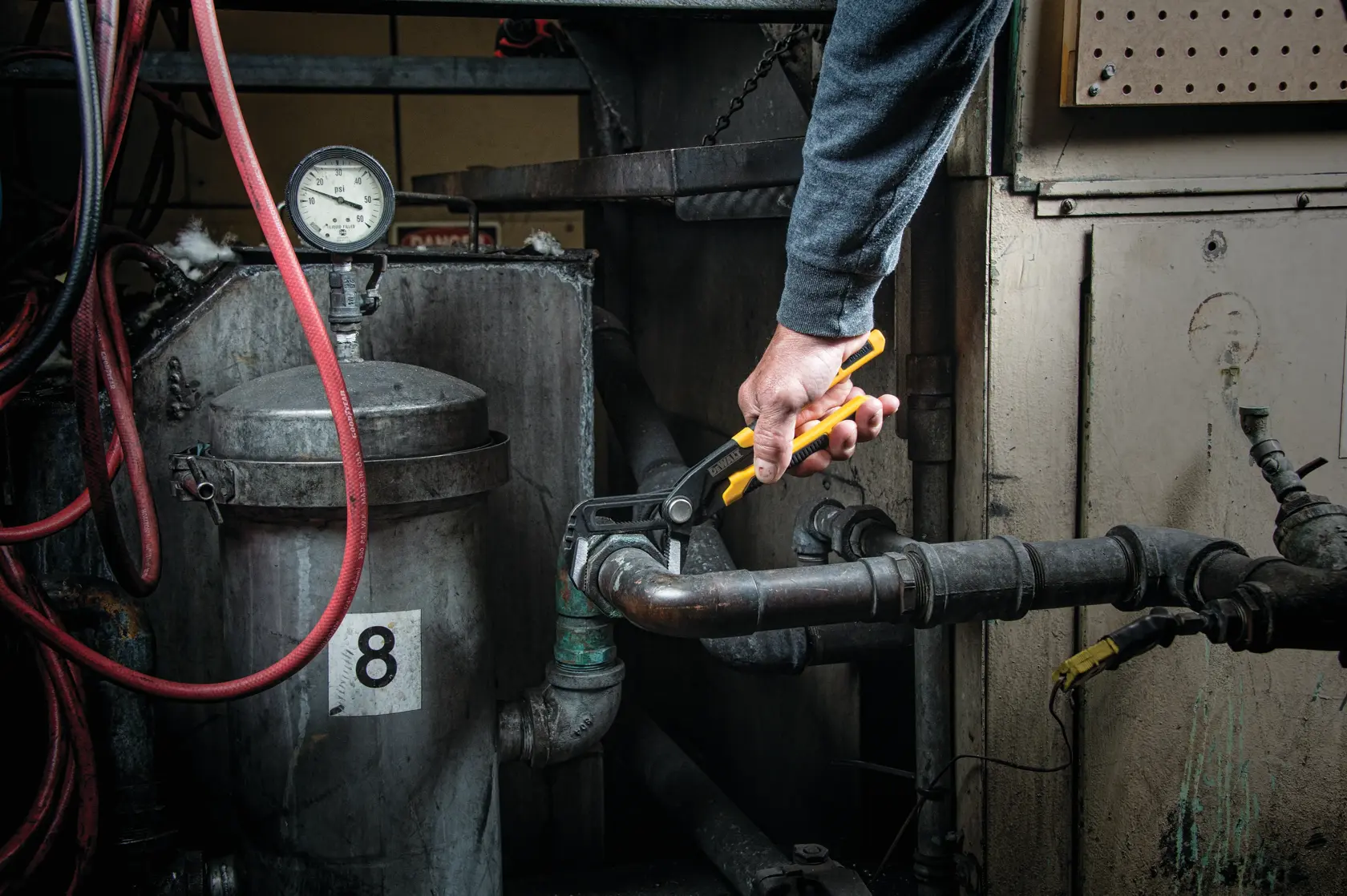 A person uses a DEWALT adjustable wrench to tighten a pipe fitting in an industrial setting with gauges and pipes.