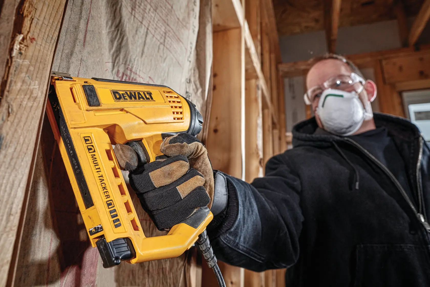 A person wearing work gloves and a mask is using a yellow DEWALT multi-tacker stapler on wooden construction framing inside a building under construction.