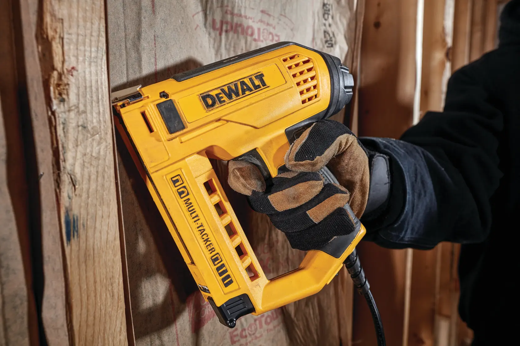 A person wearing work gloves uses a DEWALT electric staple and nail gun to fasten material onto wooden studs.