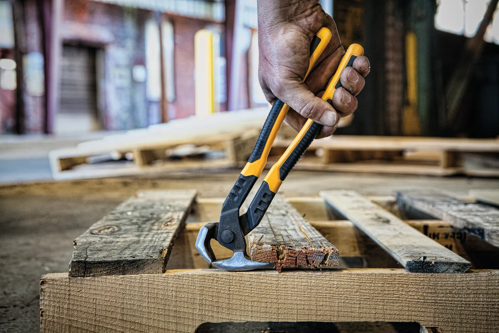 A hand using a DEWALT claw bar tool to remove a wooden board from a pallet. The tool has a black and yellow handle.