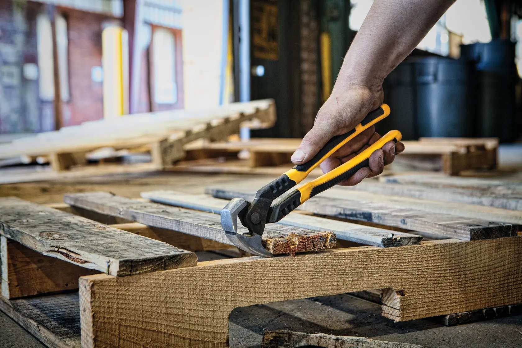 A hand uses a yellow and black DEWALT nail puller tool to extract a nail from a wooden pallet in a workshop setting.