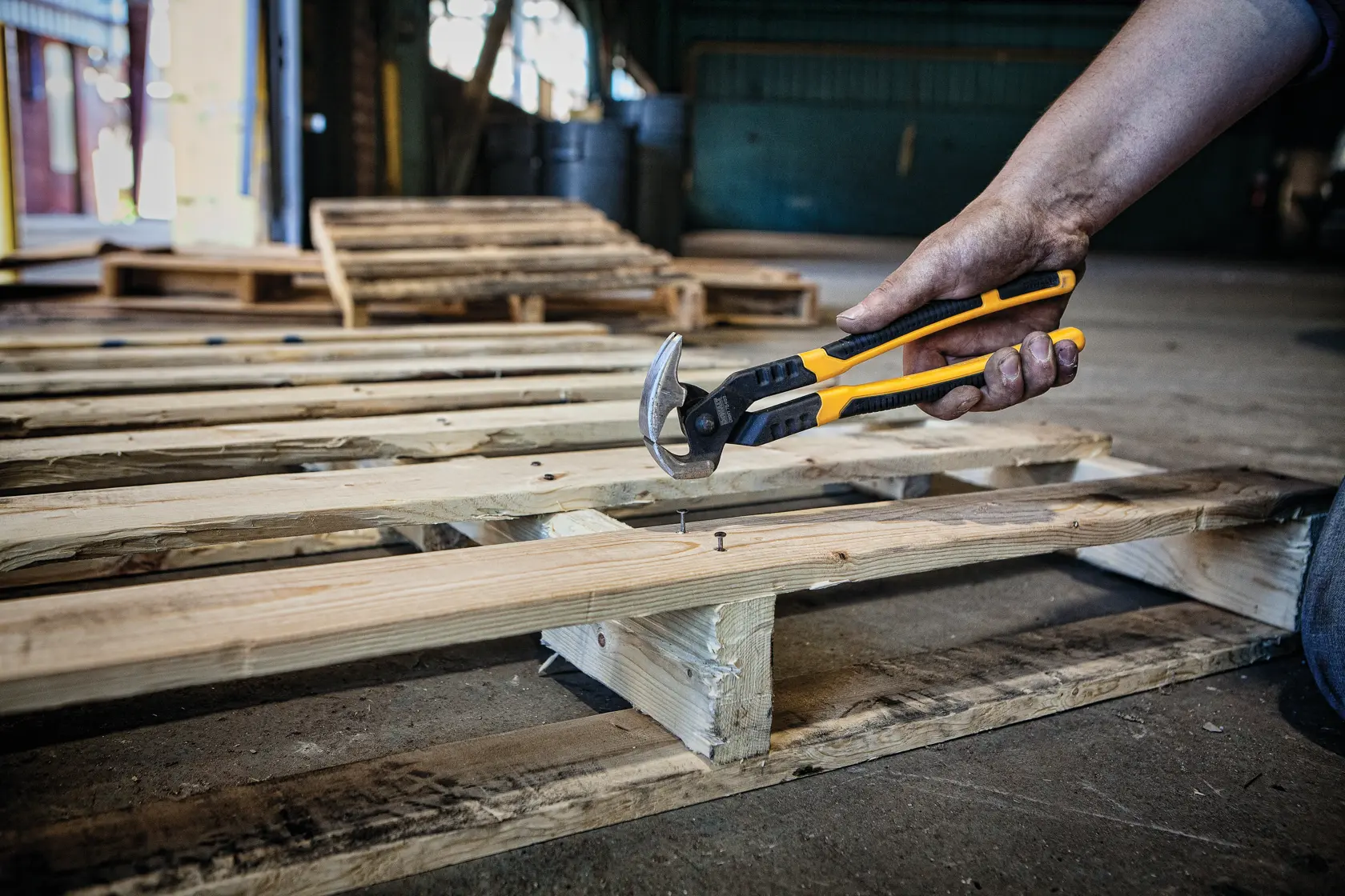 A person uses a DEWALT hand tool to remove a nail from a wooden pallet in an industrial setting.