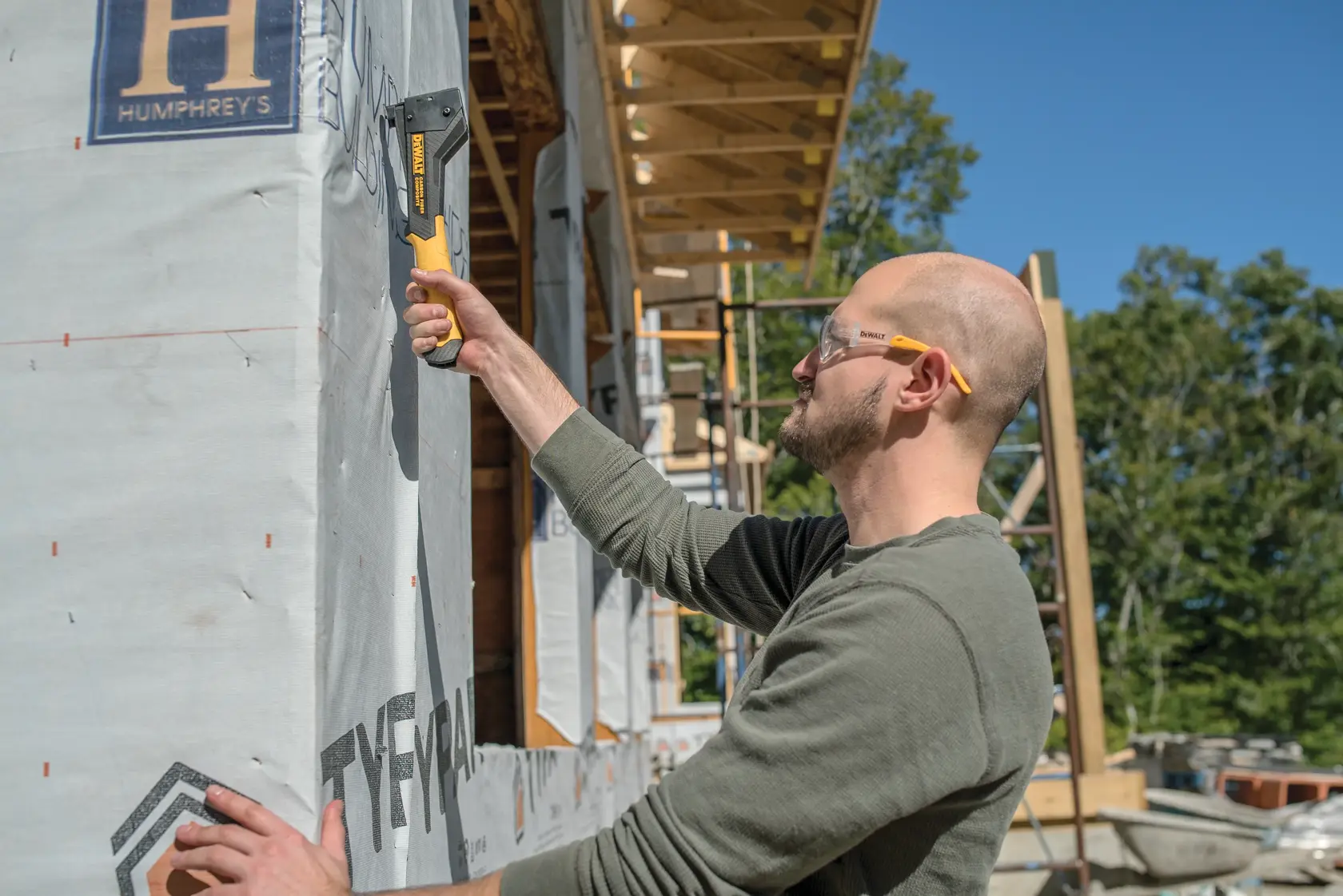A person using a DEWALT staple gun (model DWHT75900) to secure house wrap material to the exterior of a building under construction. The individual is wearing a green shirt and safety glasses, and scaffolding and trees are visible in the background.