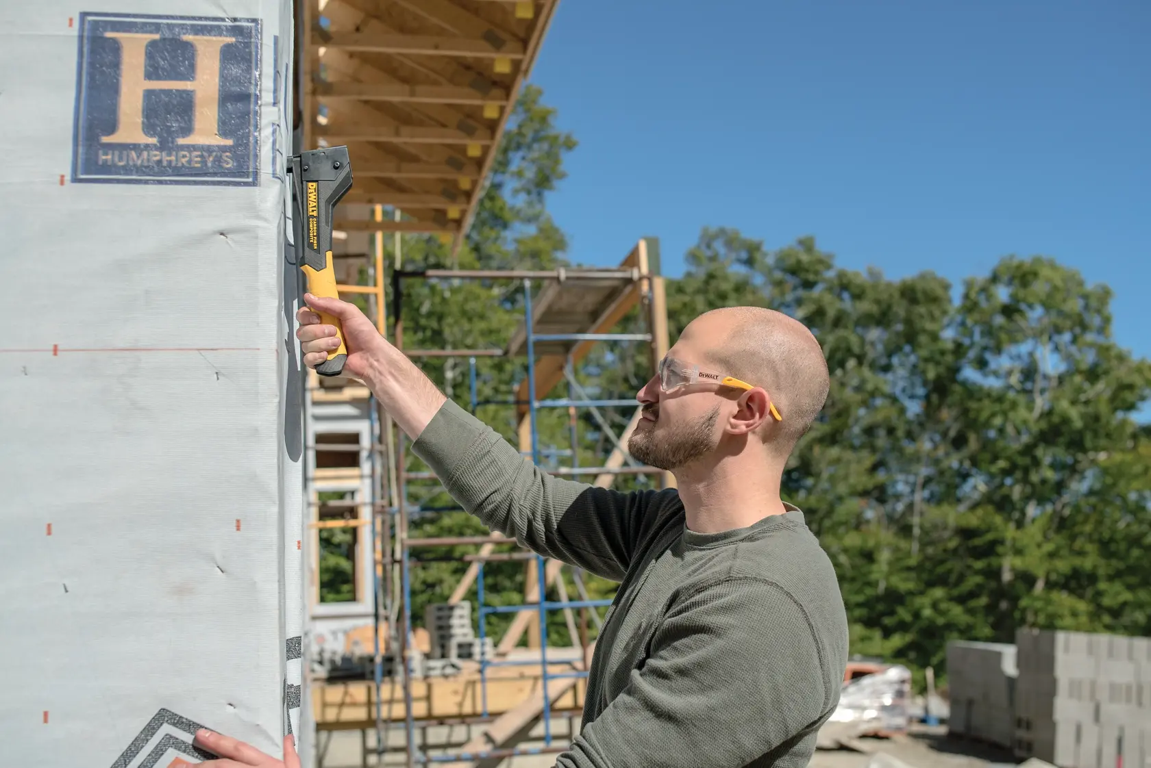 A person using a DEWALT DWHT75900 stapler to secure building wrap onto an exterior wall at a construction site. The scaffolding and materials are visible in the background, and the person's face is blurred.