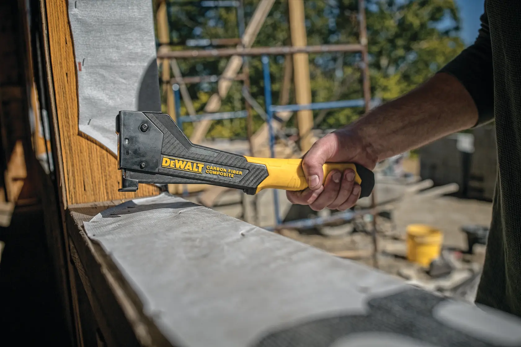A person uses a DEWALT carbon fiber composite hammer tacker to staple material onto a wooden frame at a construction site.