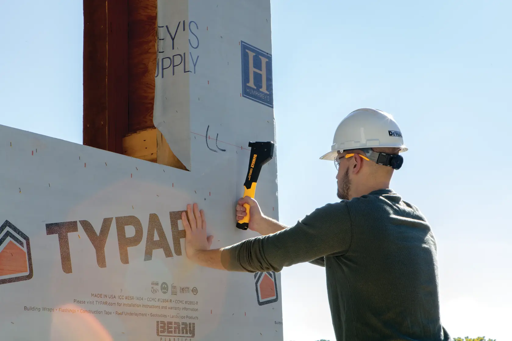 A person wearing a hard hat and safety glasses uses a DEWALT staple gun (DWHT75900) to secure building wrap to a wooden structure outdoors.