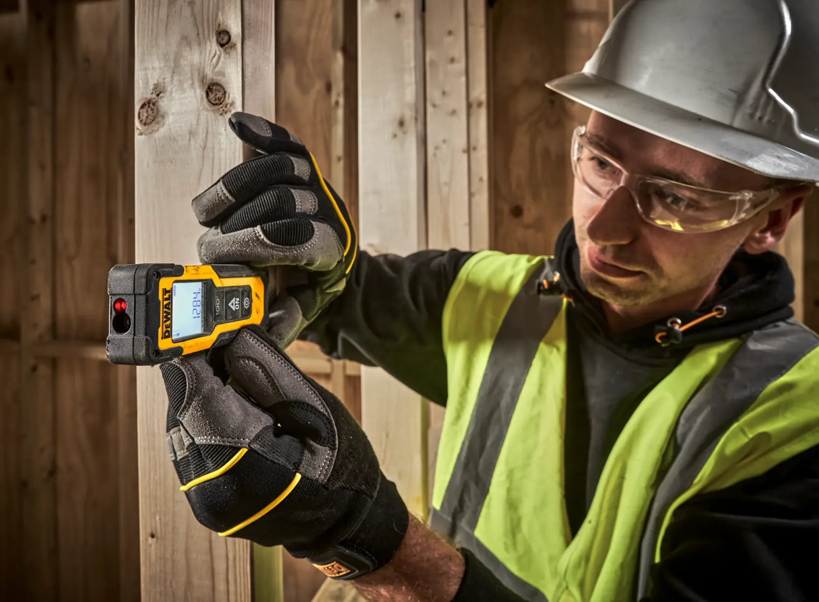 A person wearing a safety vest, work gloves, and a hard hat is using a DEWALT laser distance measurer device to measure the width of wooden planks at a construction site. The person's face is blurred for privacy.