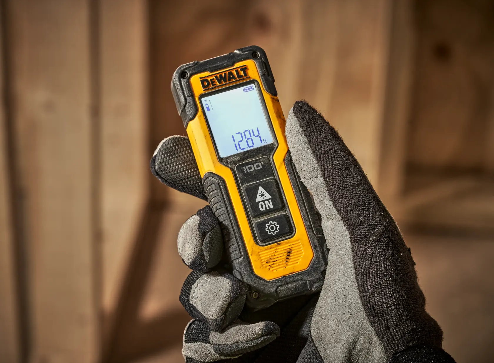 A gloved hand holding a DEWALT laser distance measurer with a digital screen displaying measurement readings, shown in a construction setting with wooden beams in the background.