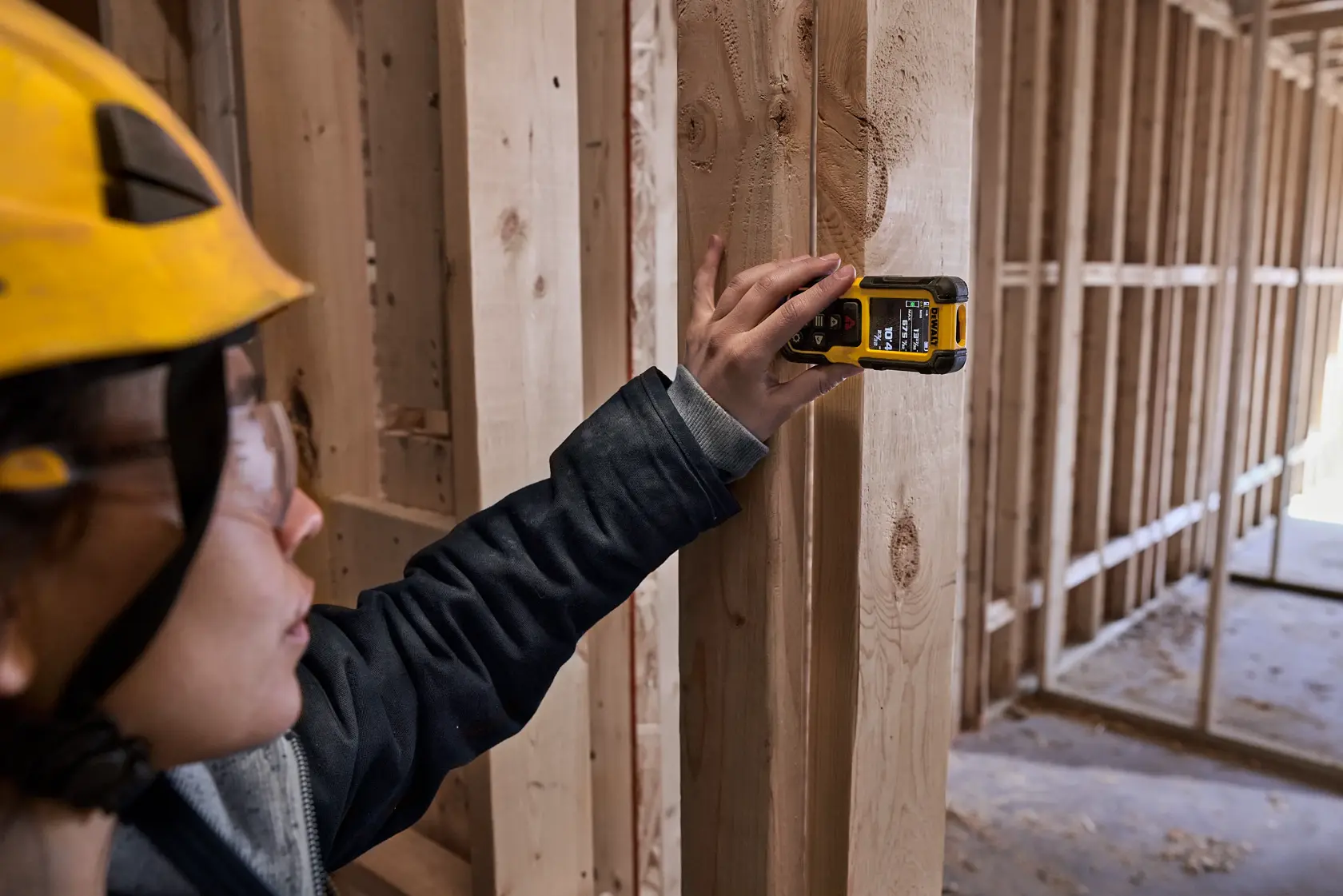 A person wearing a yellow hard hat uses a DEWALT DWHT78200 digital tool to measure or scan wooden framing at a construction site. The person's face is blurred for privacy.