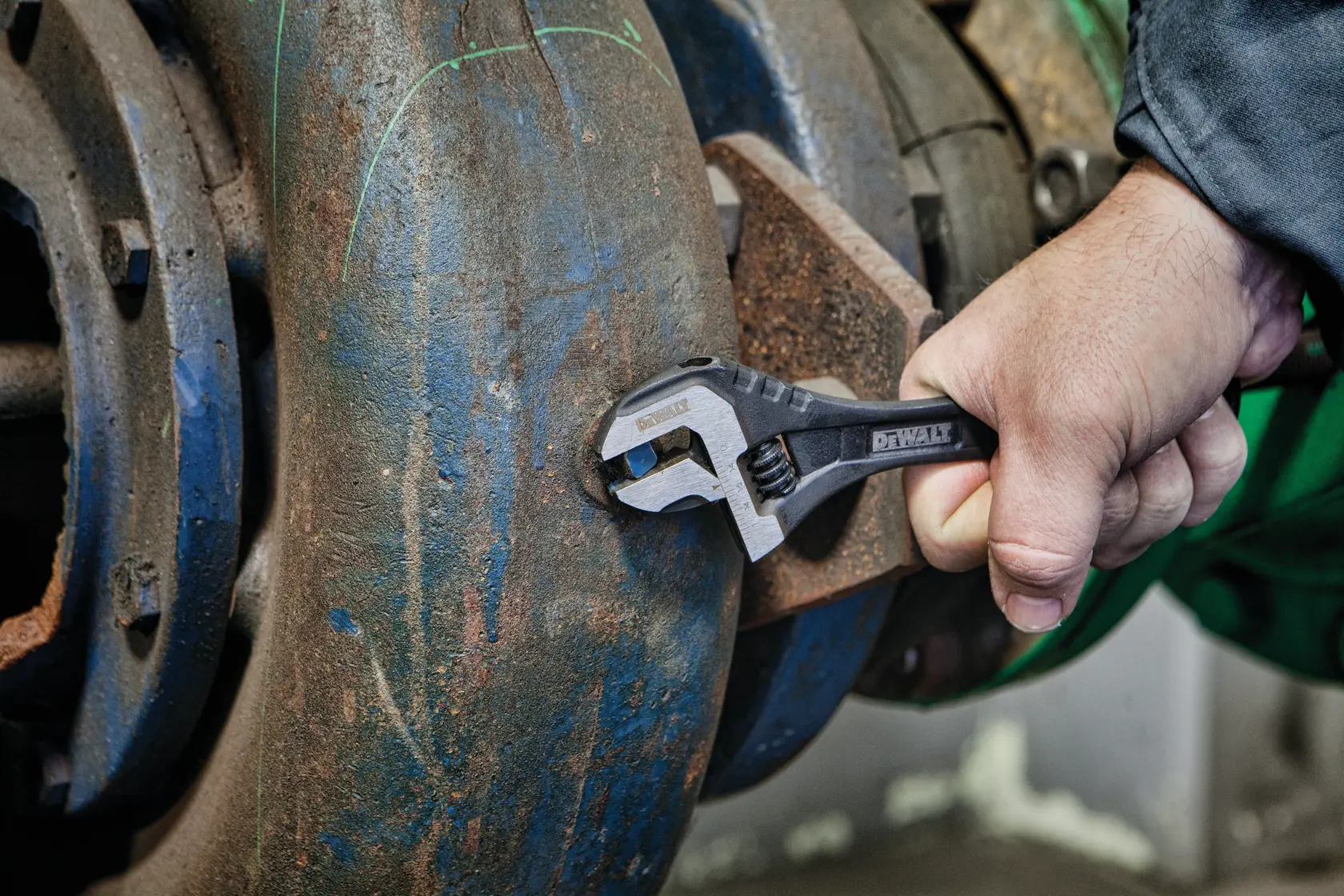 A hand using a DEWALT adjustable wrench to tighten or loosen a bolt on a large, rusted metal pipe.