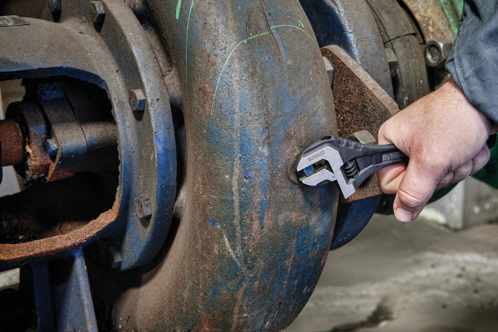 A person using a DEWALT adjustable wrench to tighten a bolt on a large, rusted industrial pipe.