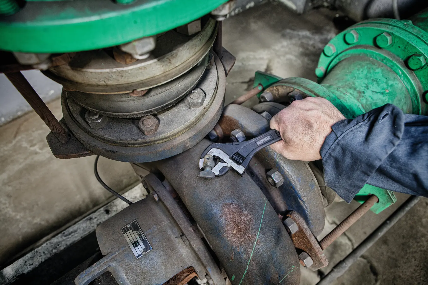 A person using a DEWALT adjustable wrench to tighten or loosen a bolt on a large industrial pipe system.