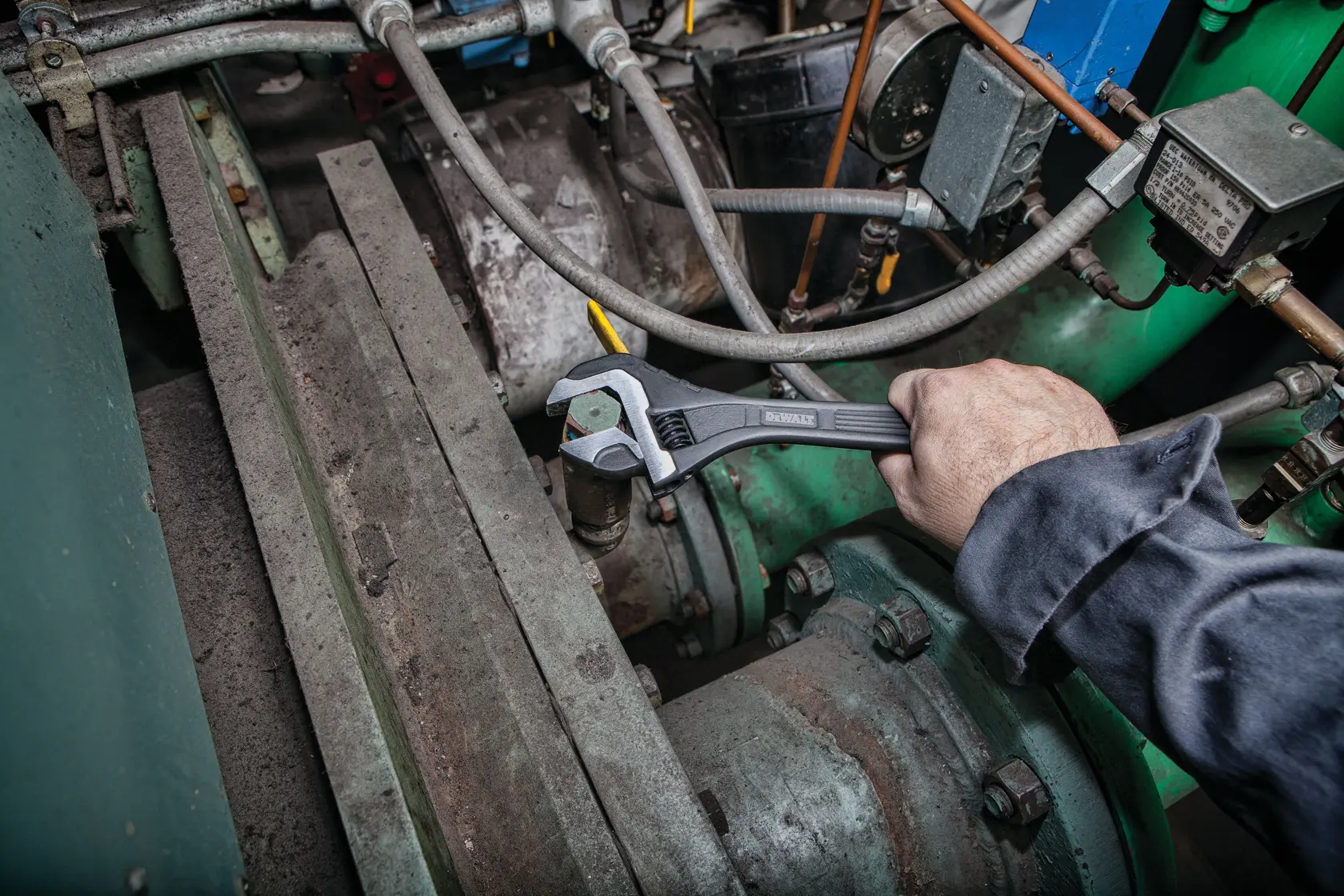 A person using a DEWALT adjustable wrench to tighten a metal bolt on industrial machinery surrounded by pipes and mechanical components.