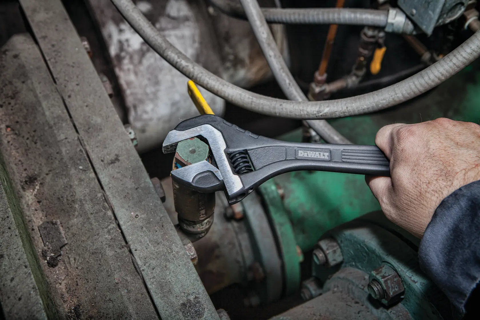 A hand using a DEWALT adjustable wrench to tighten a bolt on a mechanical part, surrounded by machinery and pipes.
