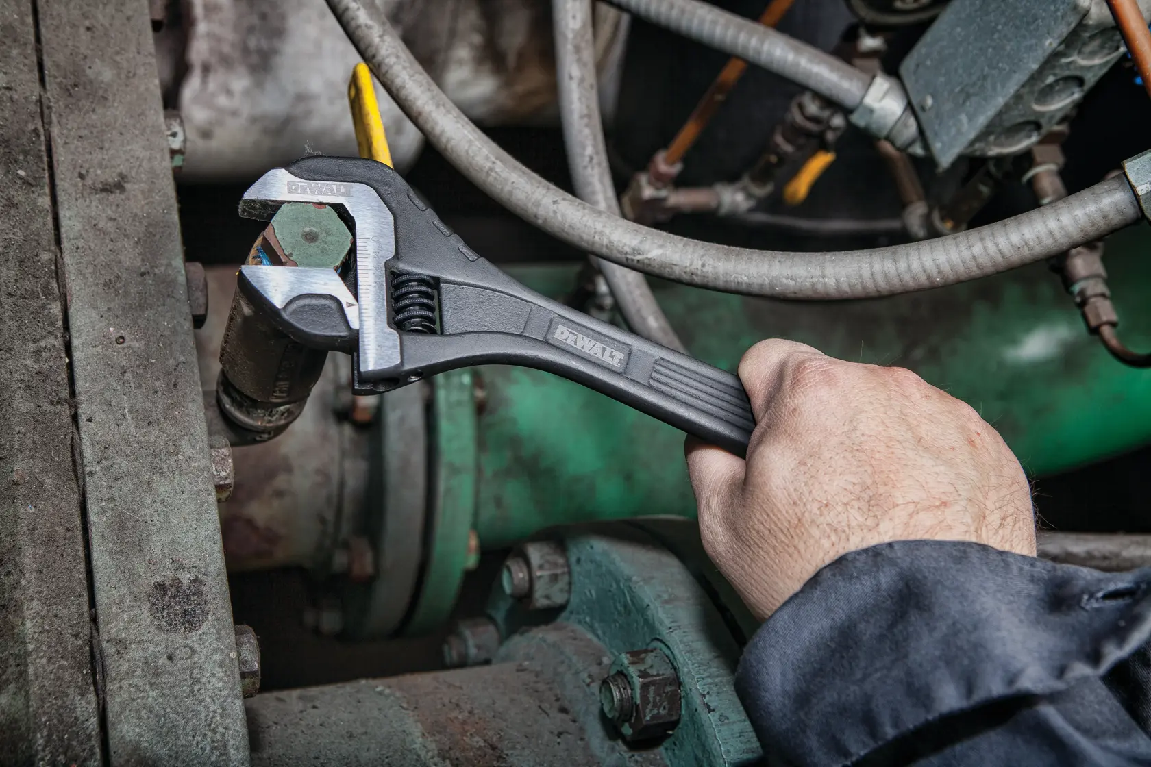 A person uses a DEWALT adjustable wrench to tighten a bolt on a piece of industrial machinery, surrounded by various pipes and mechanical components.