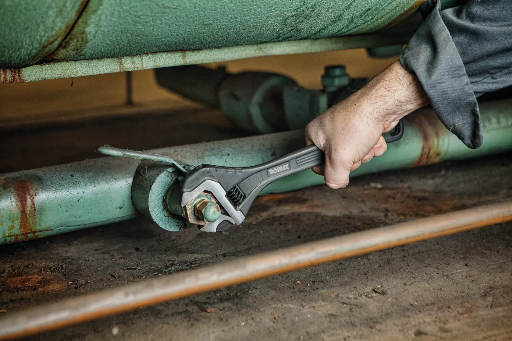 A person using a DEWALT adjustable wrench to tighten or loosen a bolt on a large, green industrial pipe.