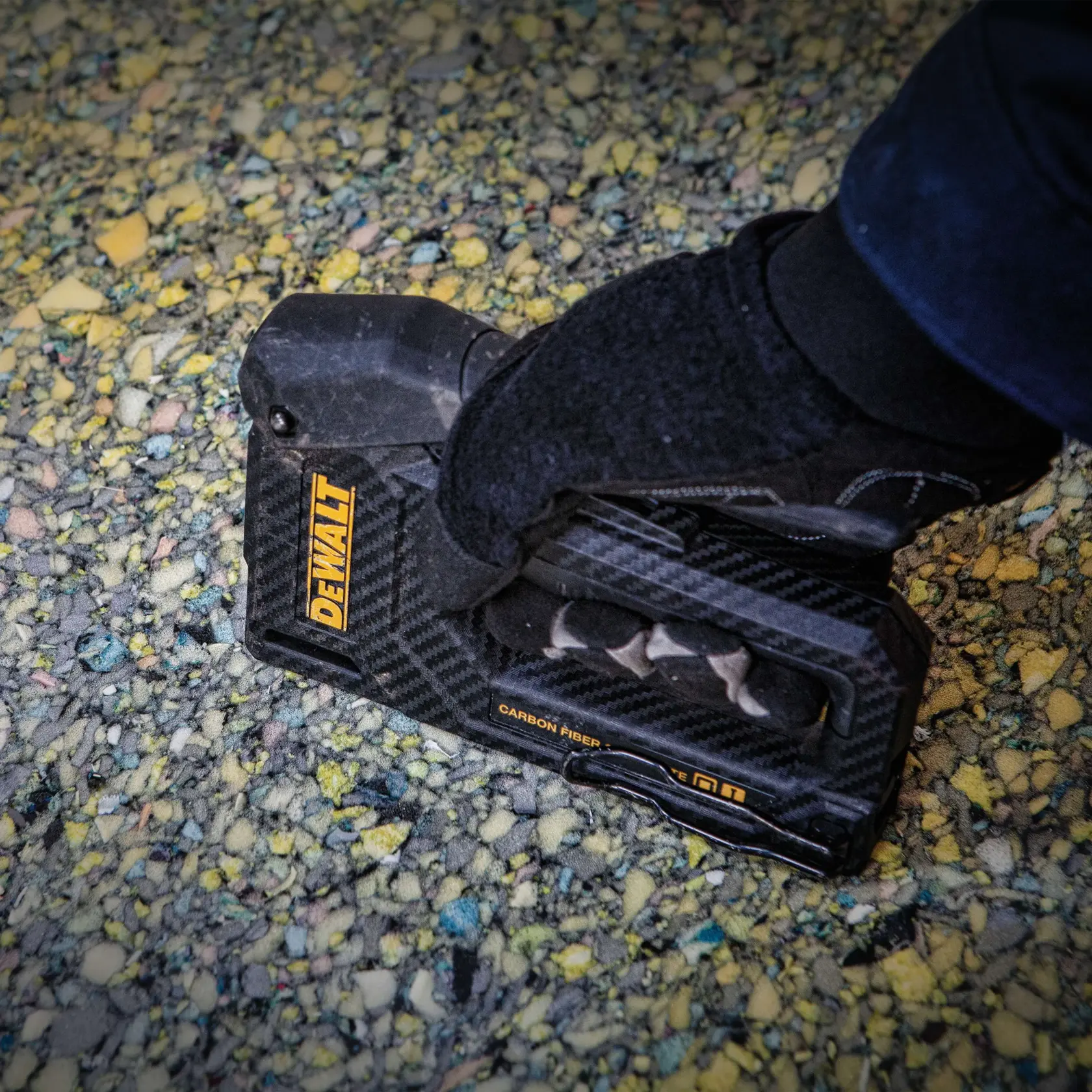 A close-up of a gloved hand holding a DEWALT carbon fiber composite hand tool, positioned on a textured surface with small colored stones.