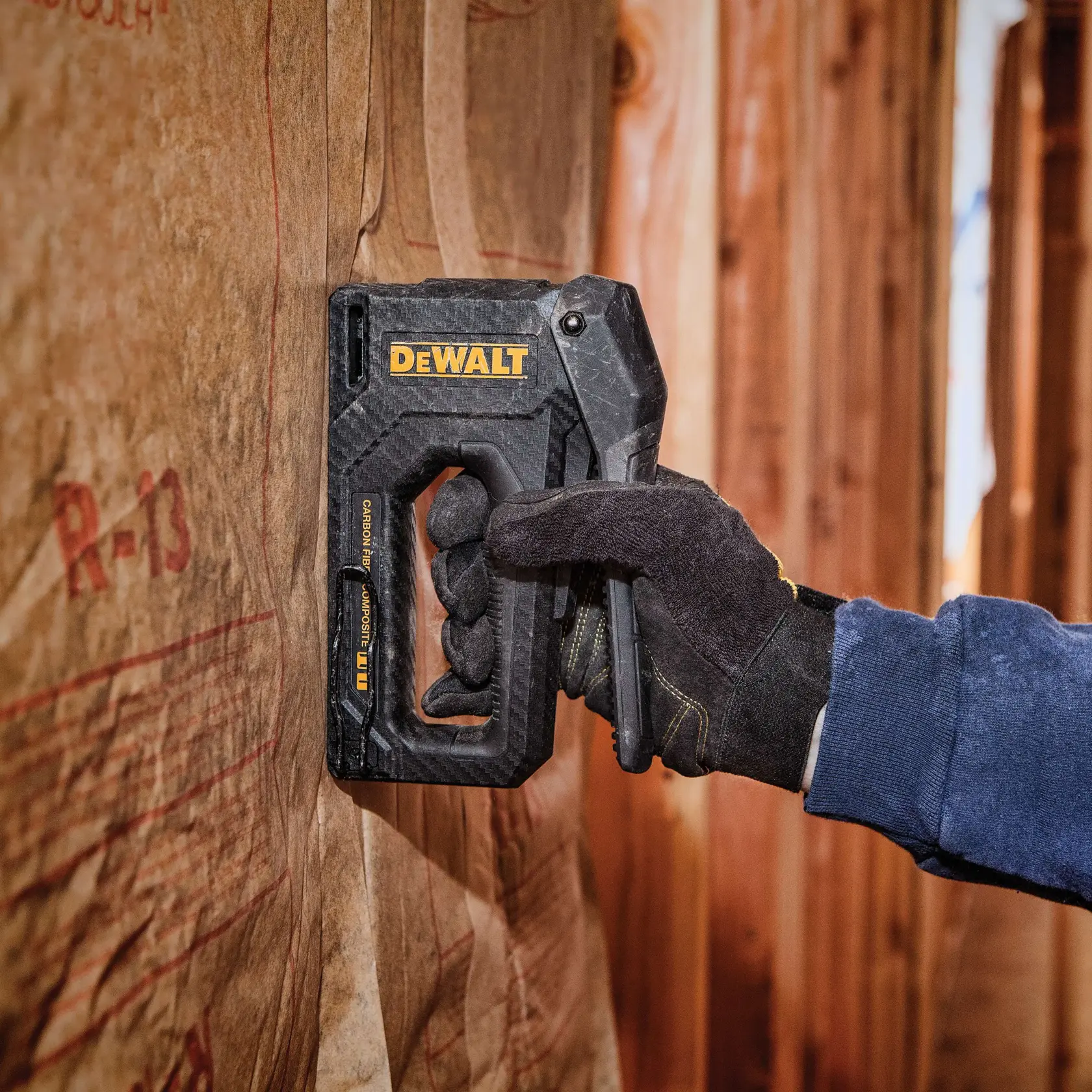 A person wearing a black work glove uses a DEWALT hand stapler to attach insulation material to a wooden wall.
