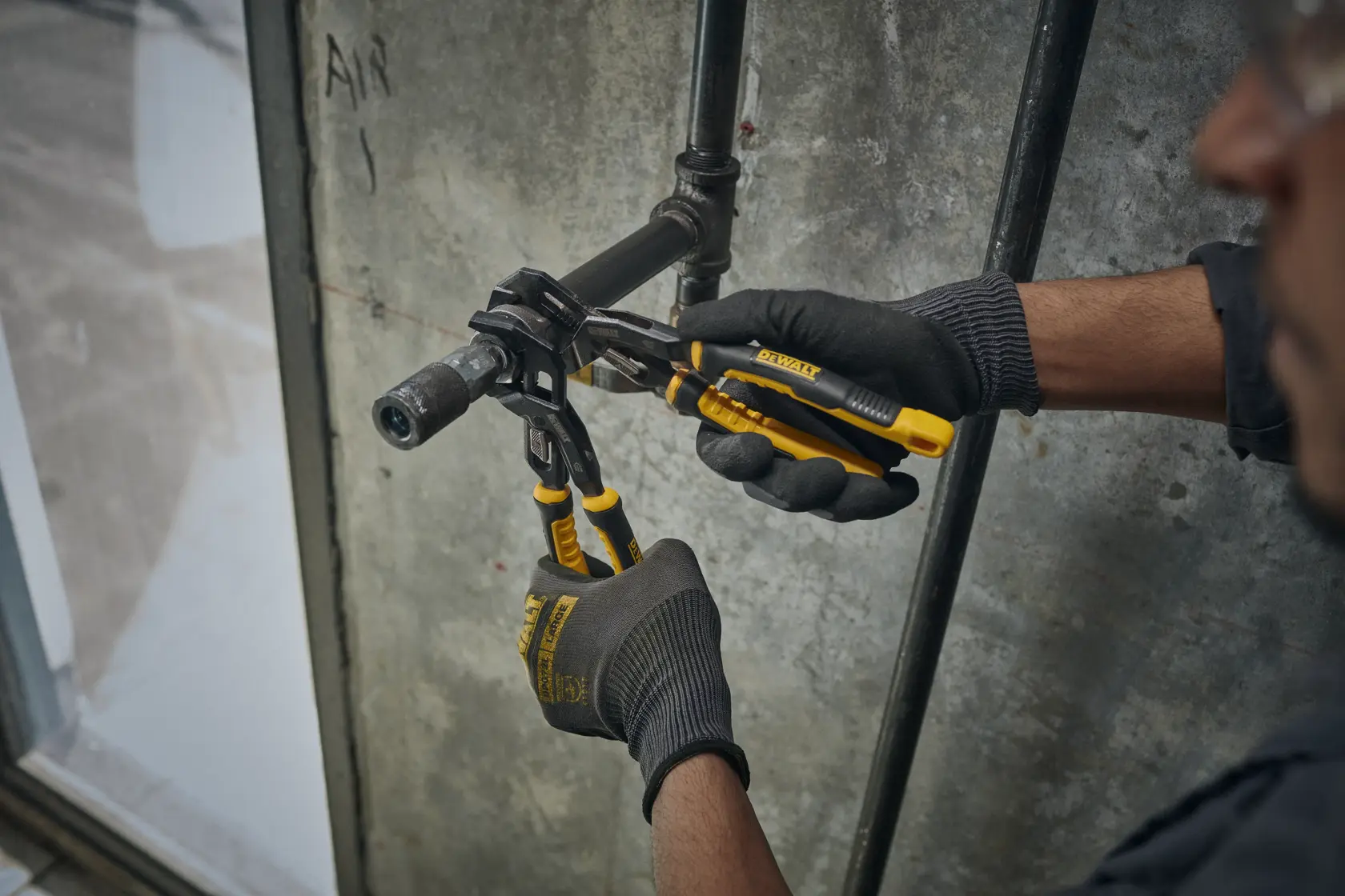 A person wearing black gloves uses DEWALT yellow and black pliers to tighten a pipe fitting against a concrete wall.