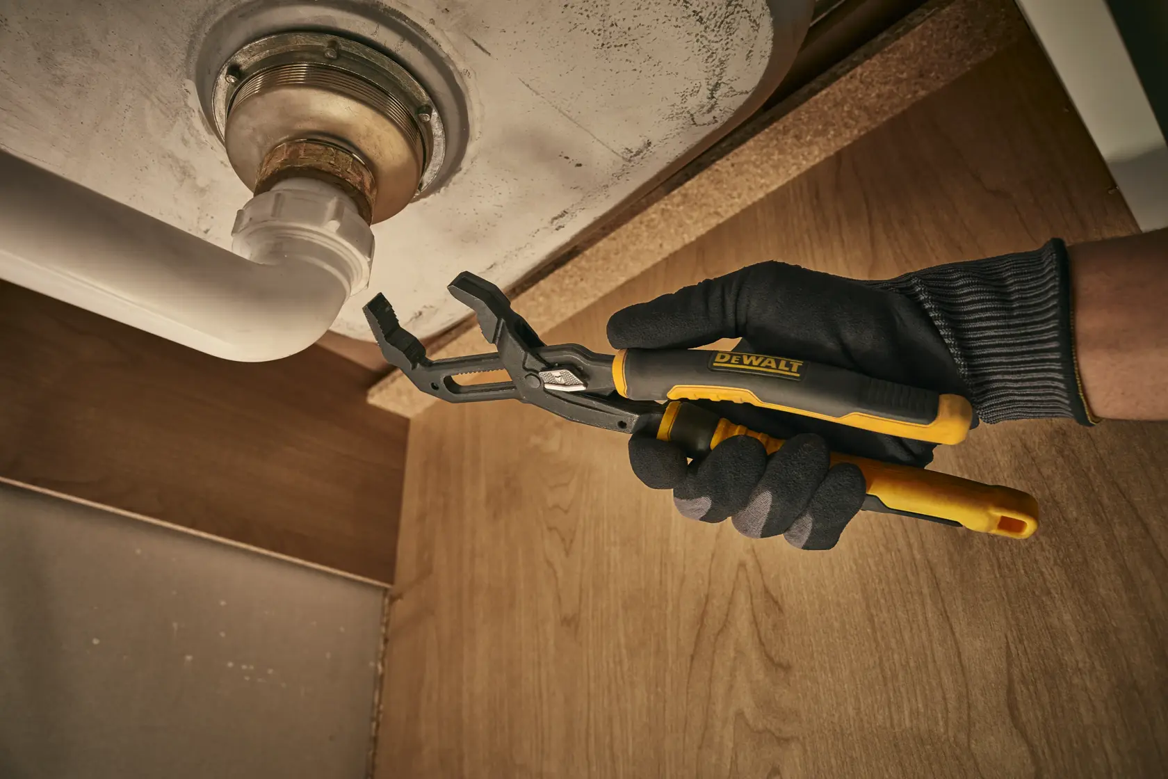 A person wearing black and gray gloves is using a yellow and black DEWALT groove joint pliers to work on a plumbing pipe under a sink.