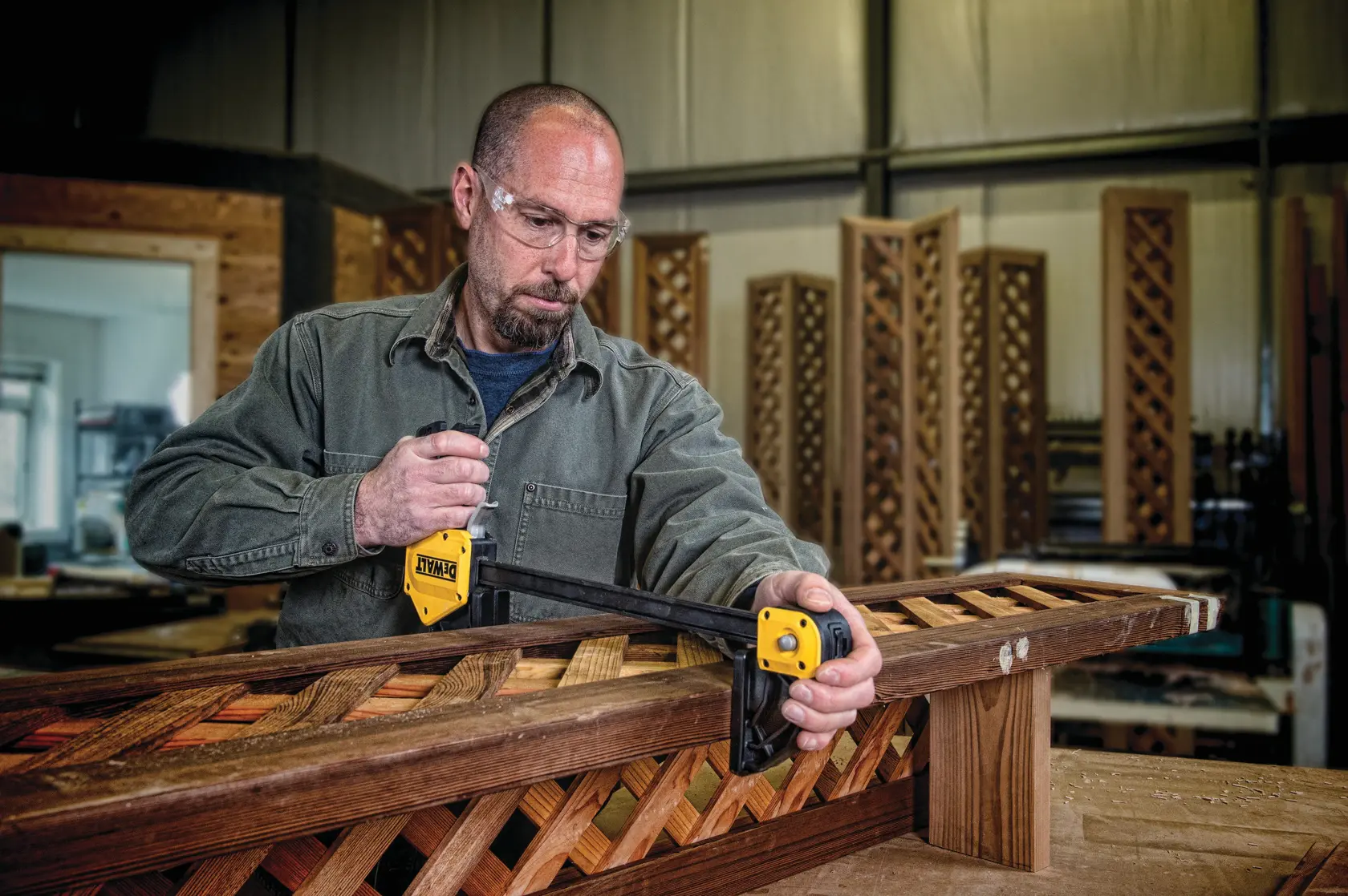 A person in a workshop uses a DEWALT DWHT83186 photography clamp to secure wooden lattice panels on a workbench. The person's face is intentionally blurred for privacy.