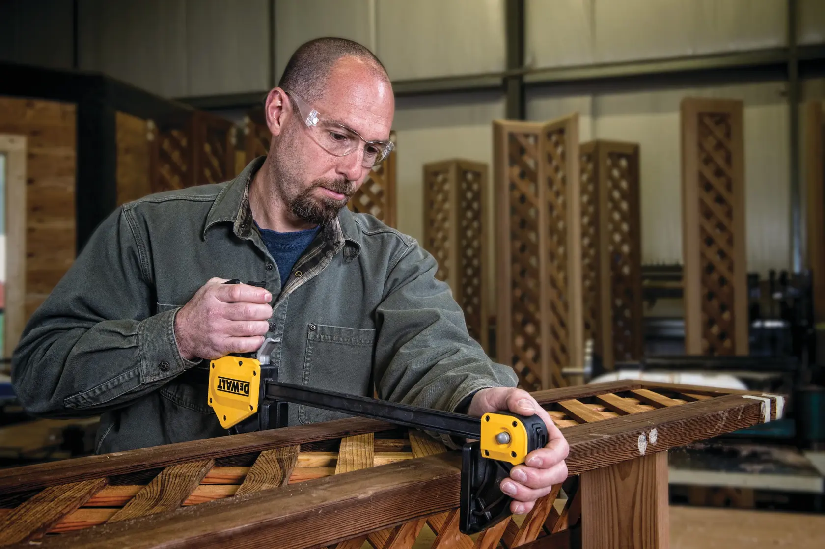 A person is using a DEWALT bar clamp (SKU: DWHT83186) to secure a piece of wooden lattice in a workshop. The individual's face is blurred for privacy.