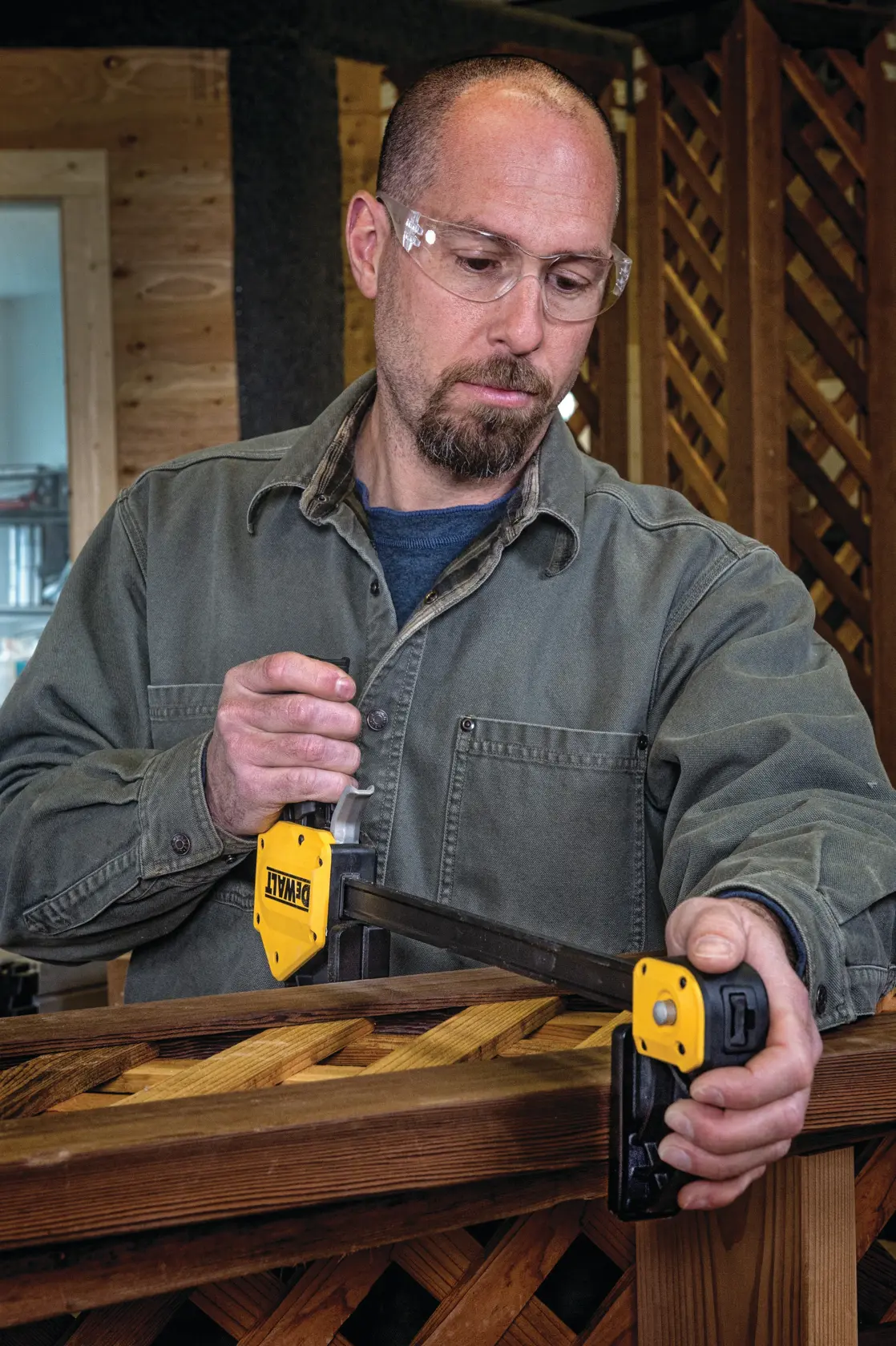 A person with their face blurred is using a DEWALT DWHT83186 woodworking clamp to secure a piece of wood with a lattice pattern, likely during a construction or carpentry project. The clamp is yellow and black and is tightly attached to the wood.