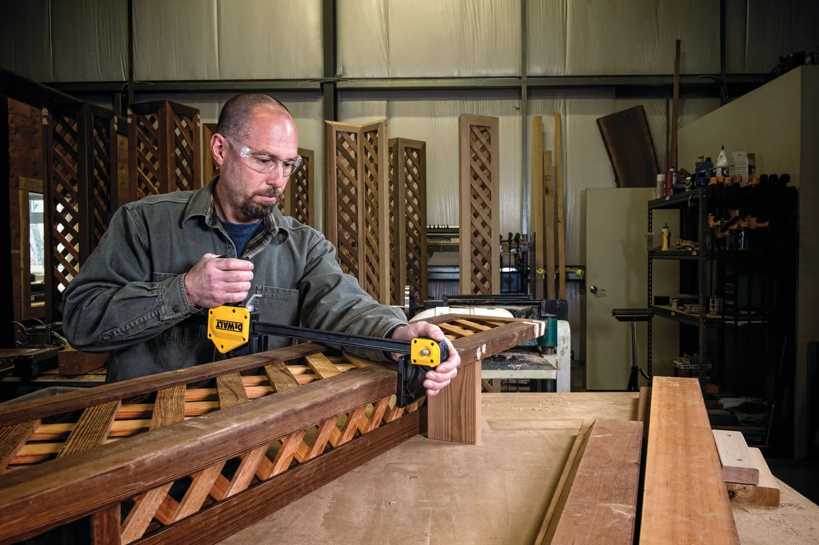 A person using a DEWALT bar clamp (model DWHT83186) to secure a wooden lattice panel on a workbench in a woodworking workshop. Various woodworking tools and materials are visible in the background.