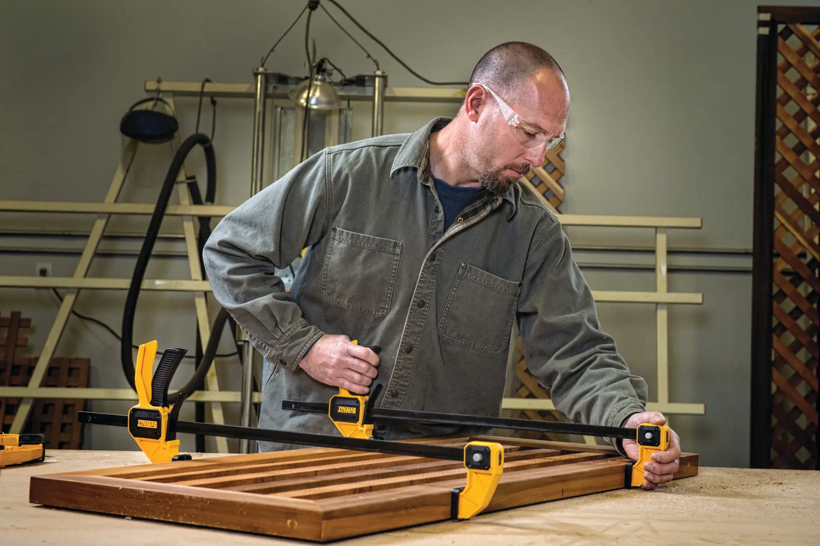 A person using yellow DEWALT clamps to secure a wooden frame on a workbench in a workshop. The person's face is blurred and the clamps are positioned at the corners of the wood.