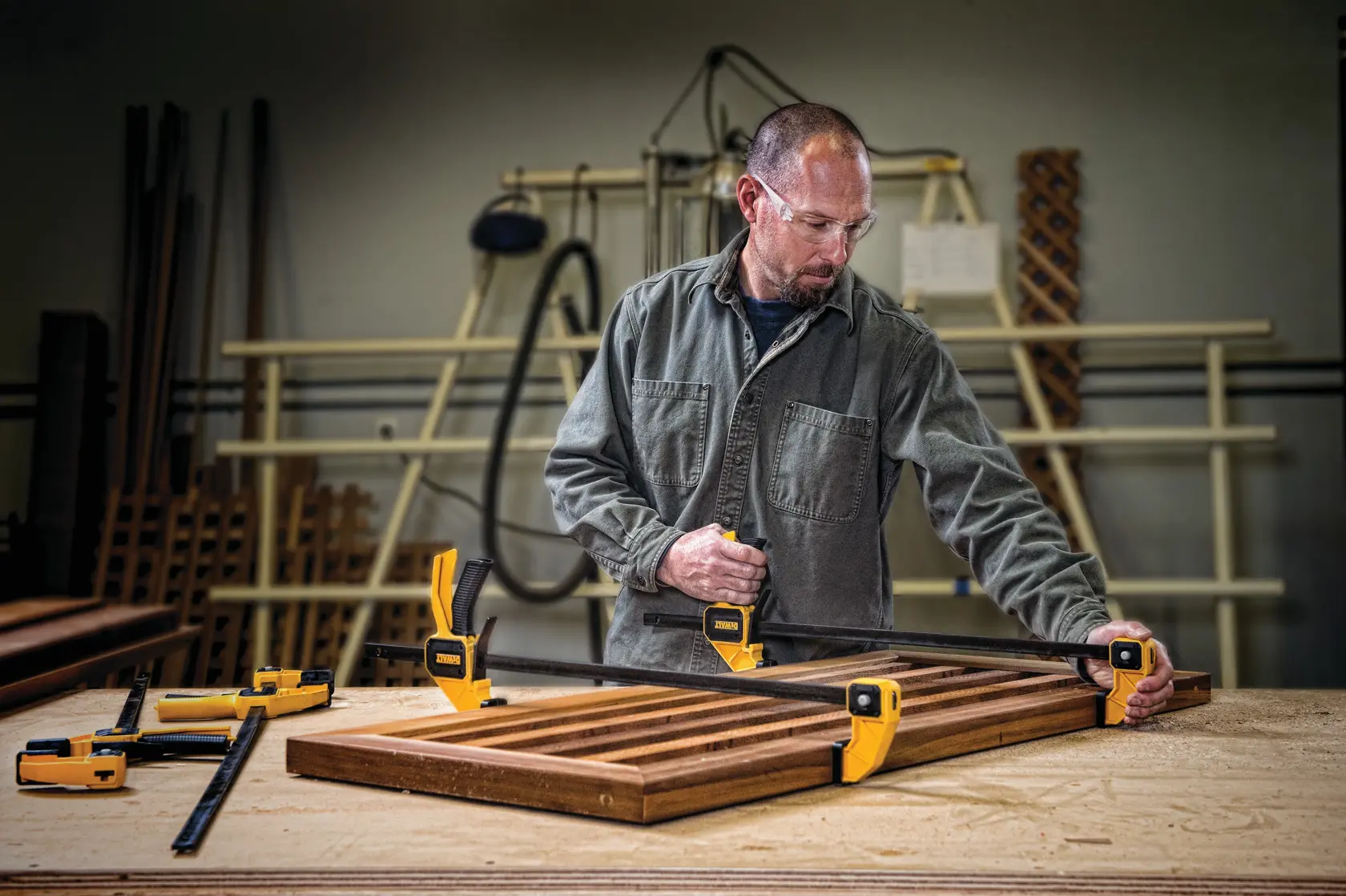 A person in a workshop uses yellow DEWALT clamps to secure a wooden frame on a workbench. Various woodworking tools and materials are visible in the background.
