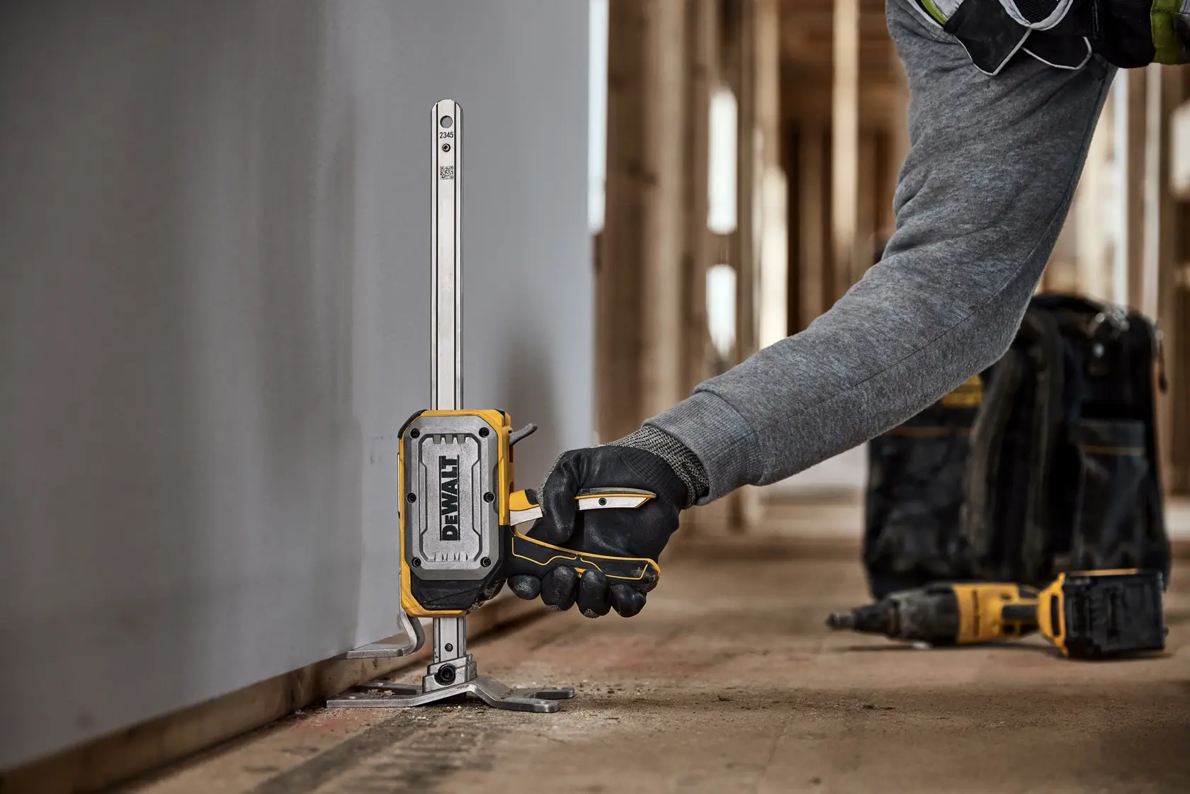 A person wearing a gray sleeve and black work gloves is using a DEWALT DWHT83550 tool to measure or align along the base of a wall at a construction site. The tool is positioned vertically against the wall, and other DEWALT equipment is visible in the background.