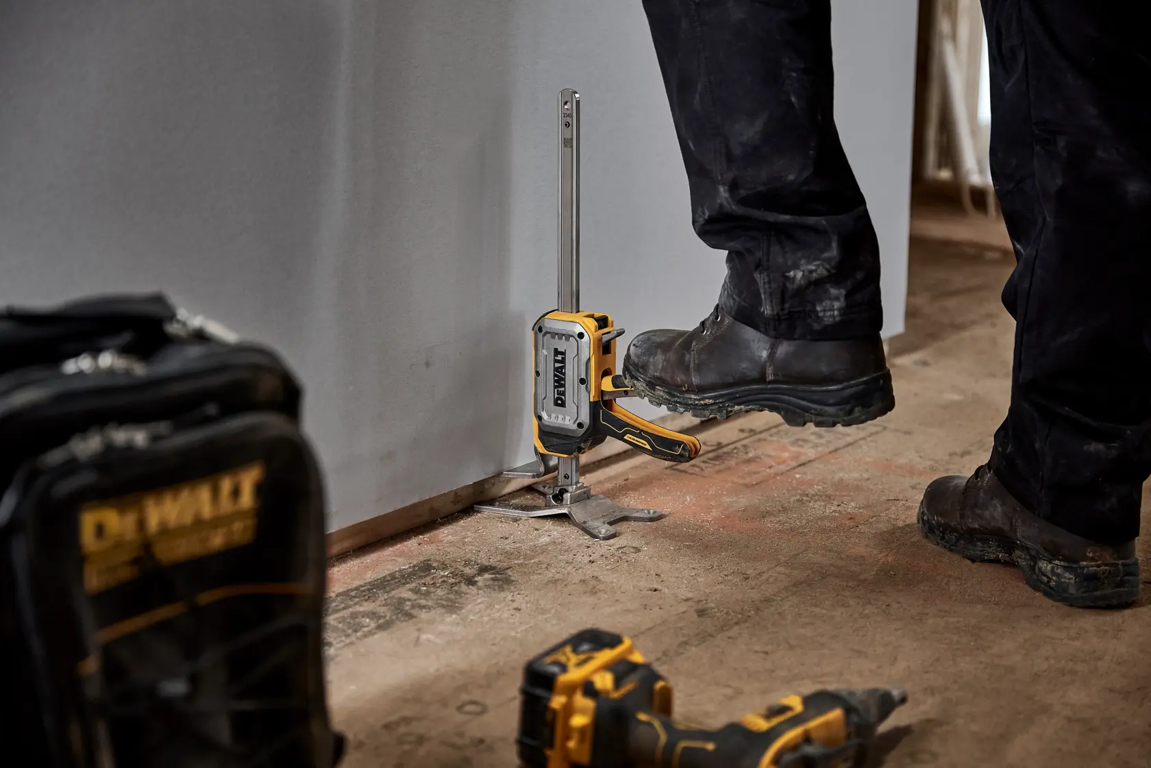 A DEWALT DWHT83550 tool is used to clamp a drywall sheet to the floor for installation. Worker wearing black pants and boots is pressing the tool in place. DEWALT bag and drill are visible on the floor.