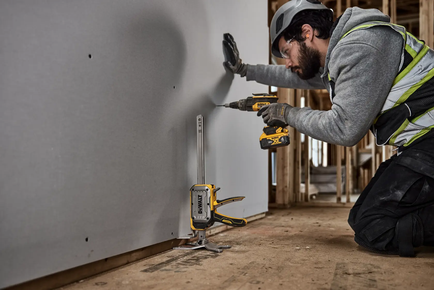 A worker wearing safety gear installs drywall using a DEWALT tool, with the DEWALT DWHT83550 clamp and guide positioned at the bottom for alignment. The worker is using a DEWALT drill to secure the panel inside a building under construction.