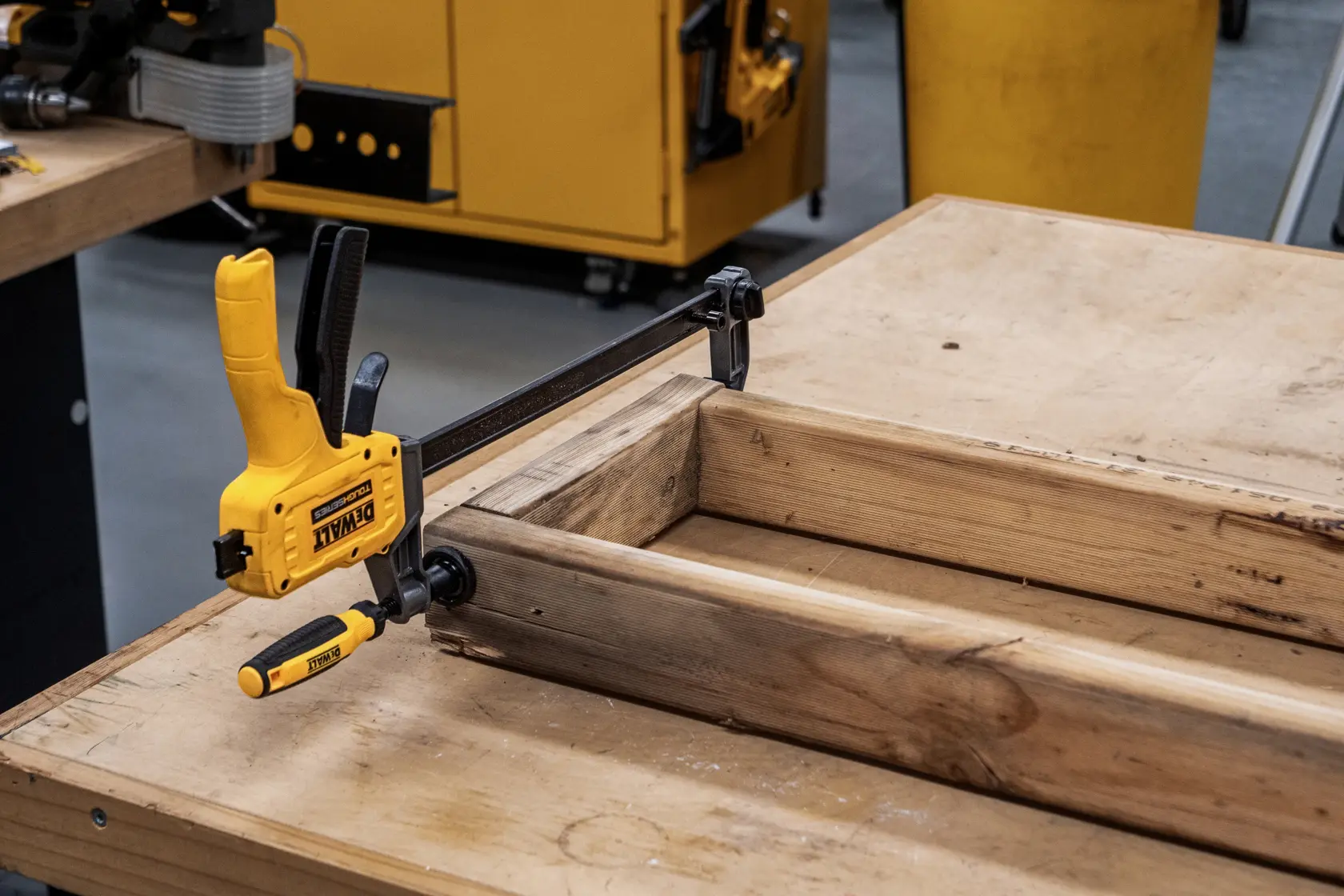 A DEWALT bar clamp, model DWHT83600, is holding pieces of wood together on a workbench in a workshop setting. The clamp is yellow and black, and is being used for woodworking or assembly tasks.