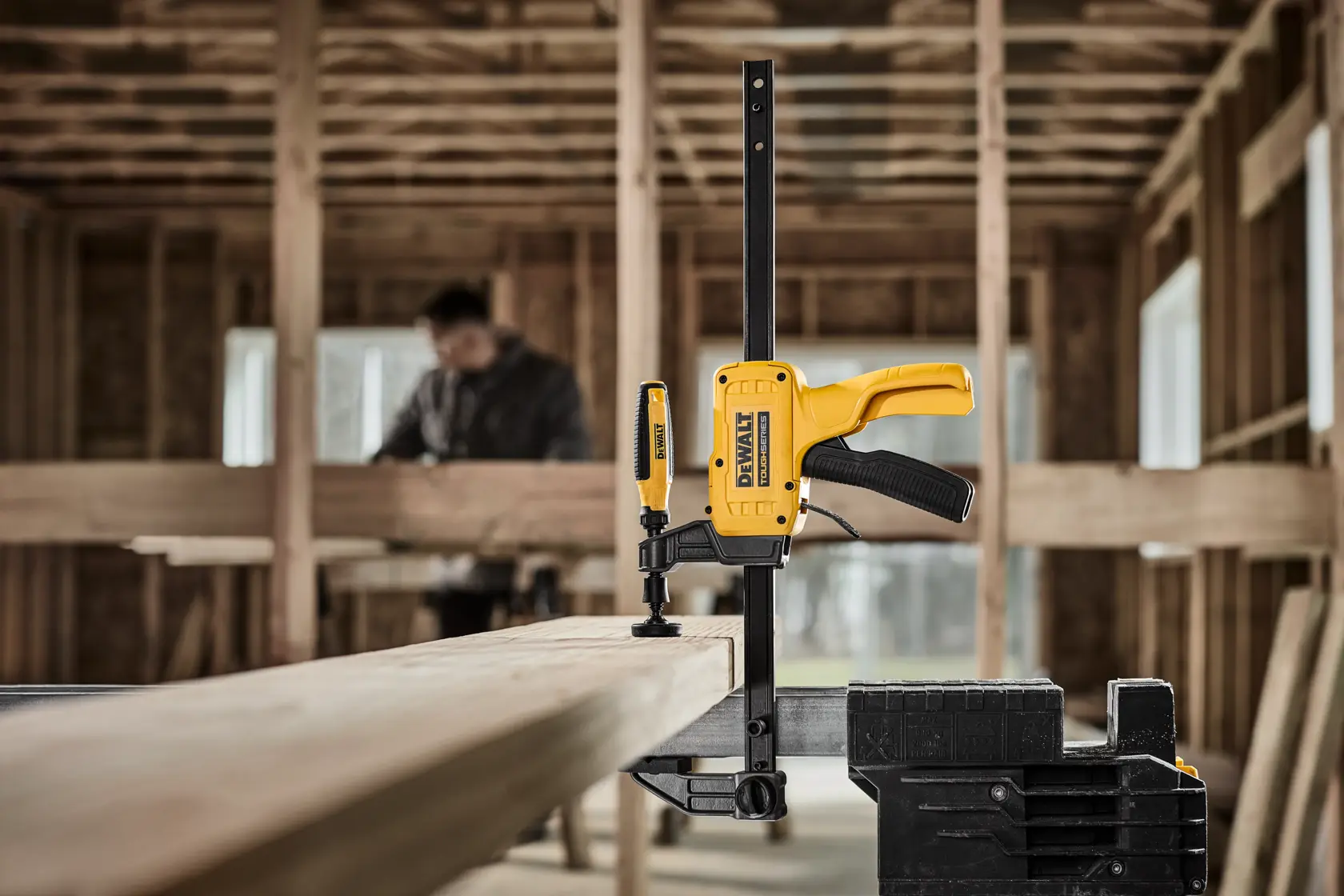 A DEWALT DWHT83600 bar clamp is securing a wooden plank in a construction environment. The yellow and black clamp is attached to a workbench, with a blurred person in the background working inside an unfinished building.